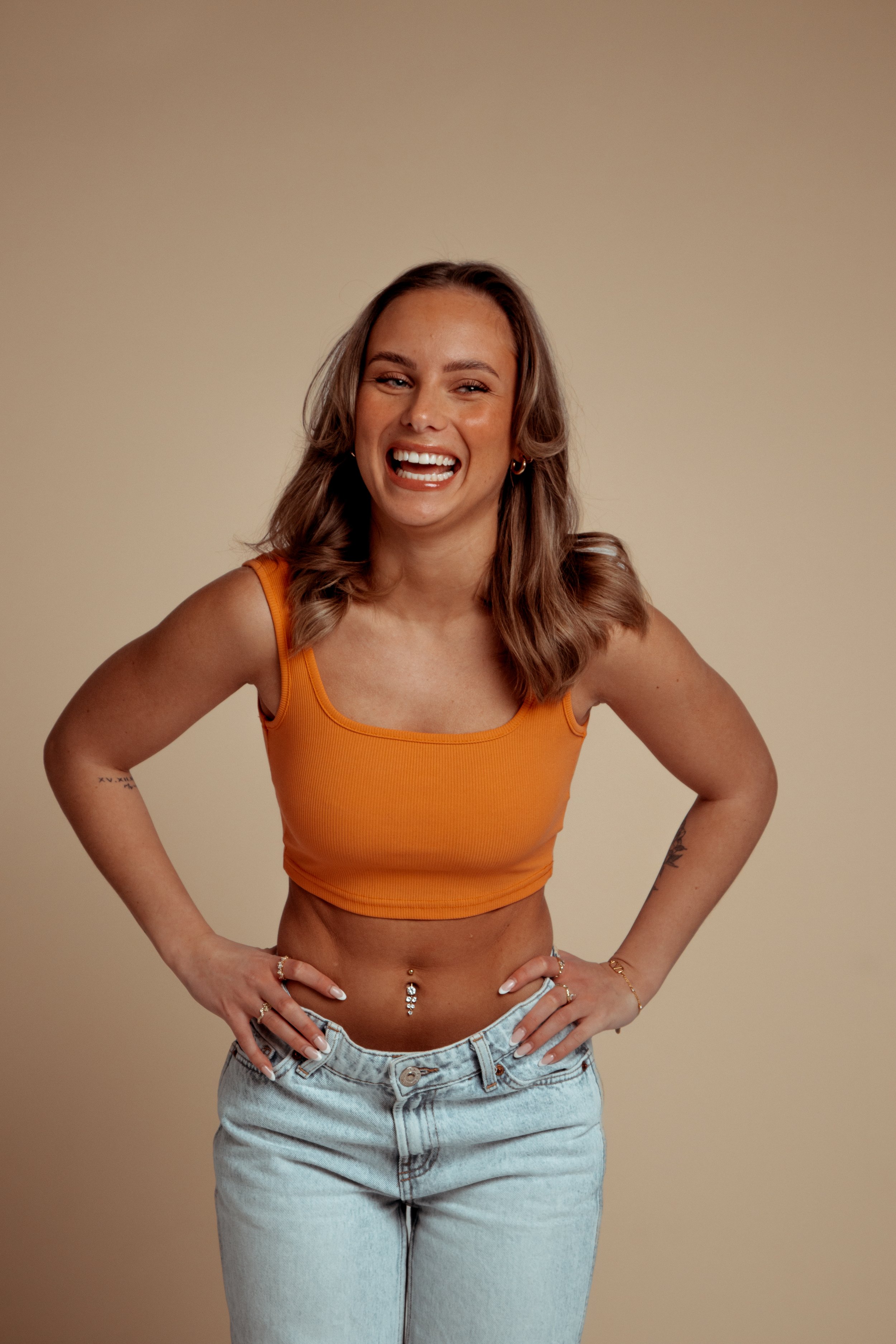 Woman with shoulder-length wavy hair, wearing a sleeveless orange crop top and light-wash jeans, smiling and posing with her hands on her hips against a plain beige background.