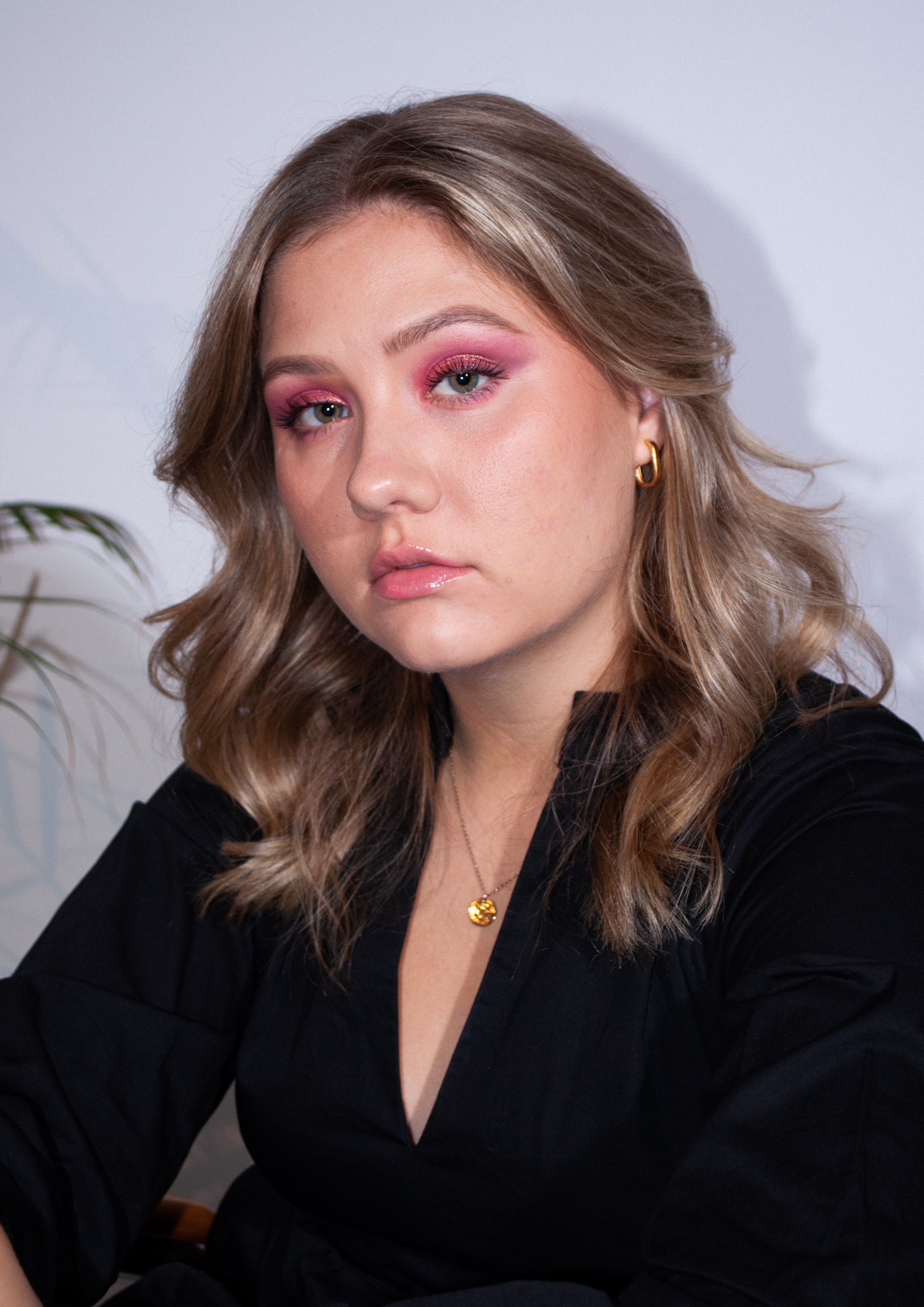 A young woman with shoulder-length wavy blonde hair, pink eye makeup, and light pink lipstick. She is wearing a black top, gold hoop earrings, and a gold necklace with a pendant. She is sitting indoors against a light-colored background with some pla