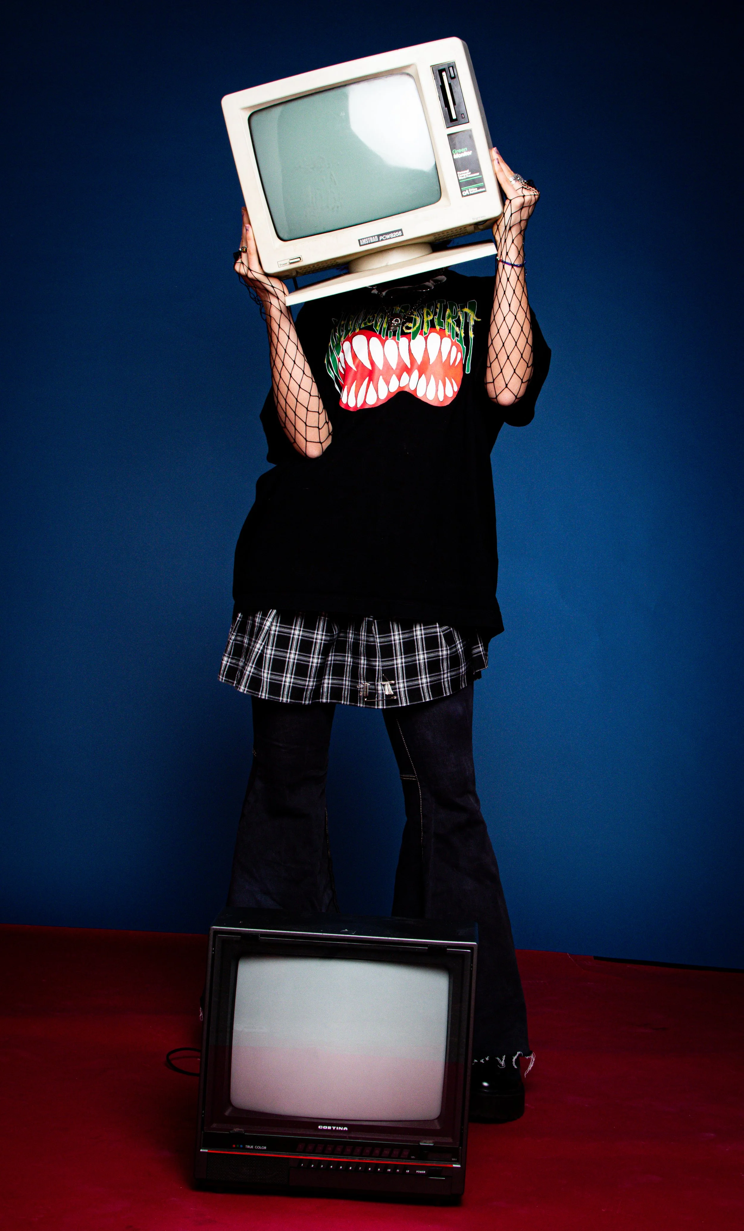 A person wearing a black shirt with a graphic monster face, fishnet sleeves, and a plaid skirt, holding an old computer monitor over their head. They are standing on a red floor with a second vintage TV on the ground, against a dark blue background.