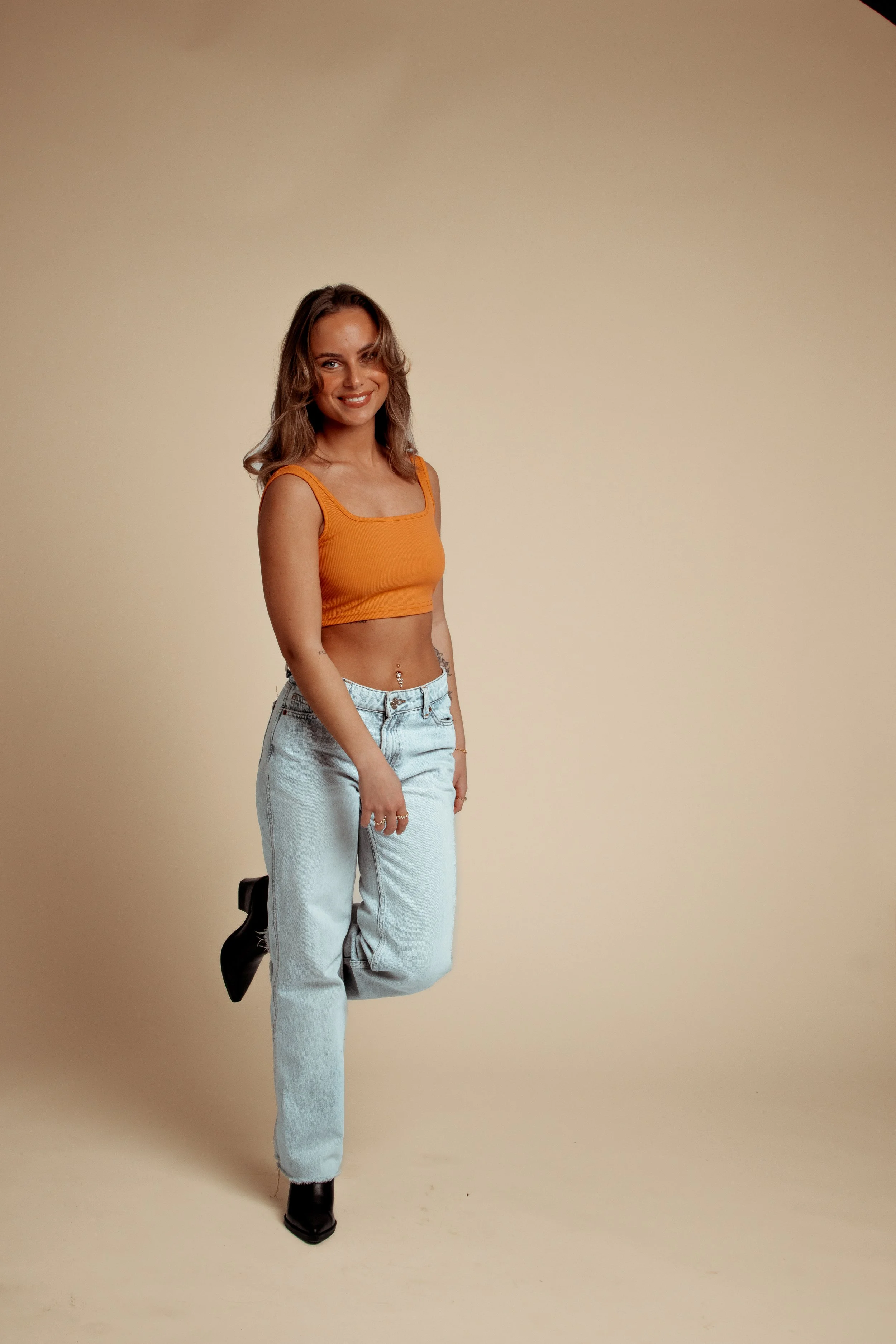 A young woman with shoulder-length wavy hair, wearing an orange crop top, light blue jeans, black boots, and standing against a beige background, smiling at the camera.