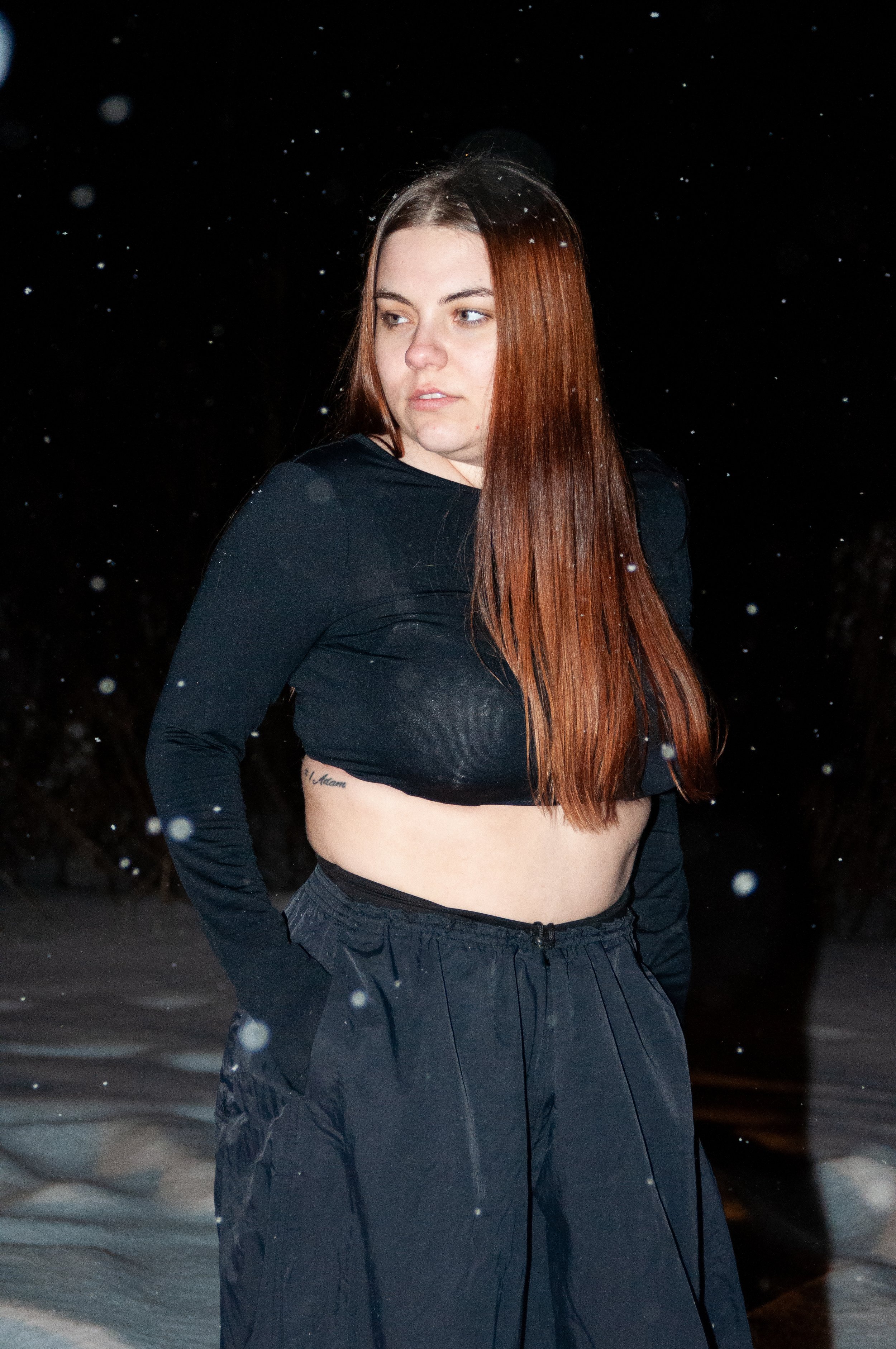 A young woman with long red hair stands outside in the snow at night, wearing a black long-sleeve crop top and black pants.