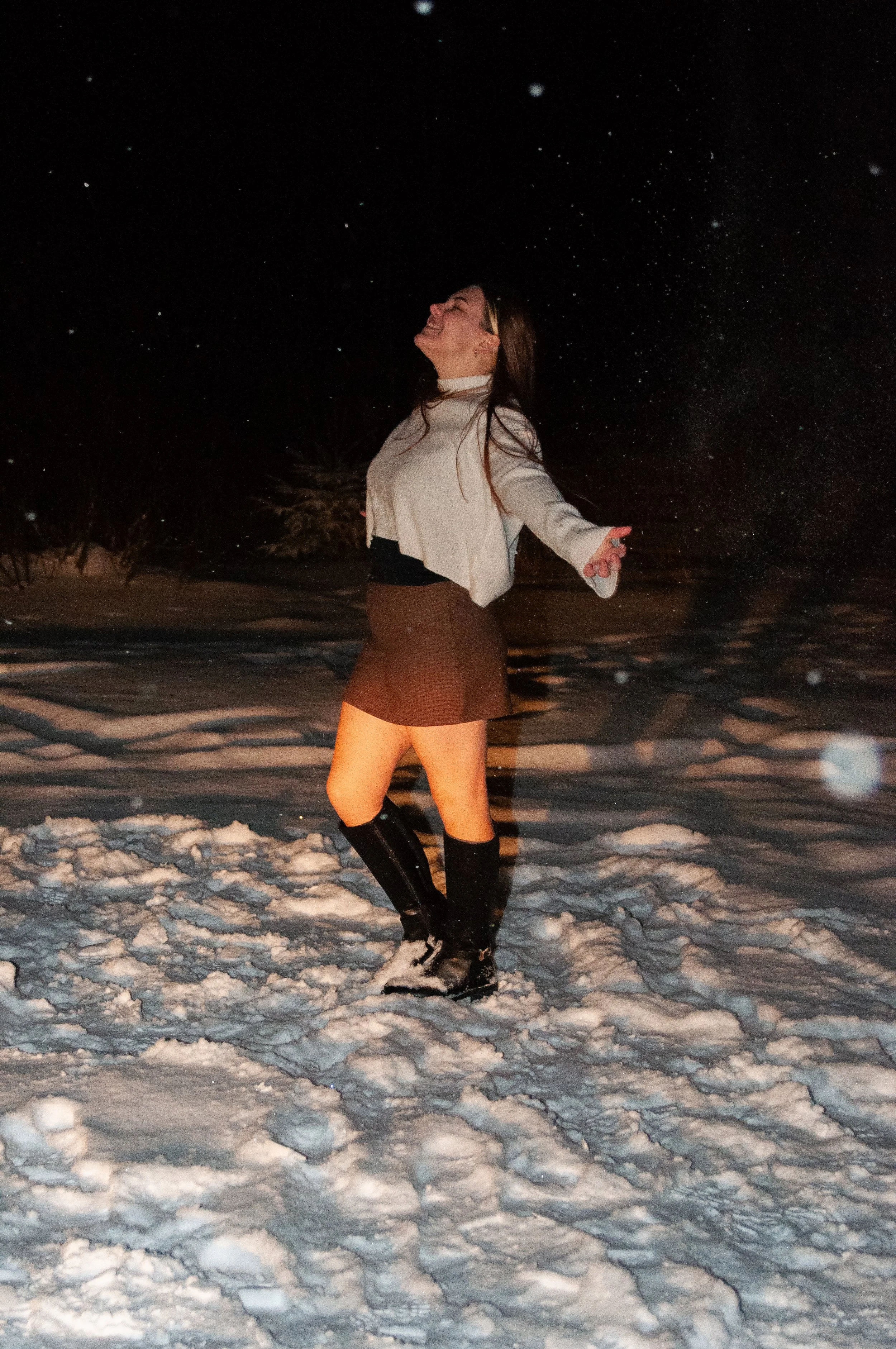 A woman standing in the snow at night with her arms outstretched and eyes closed, smiling.