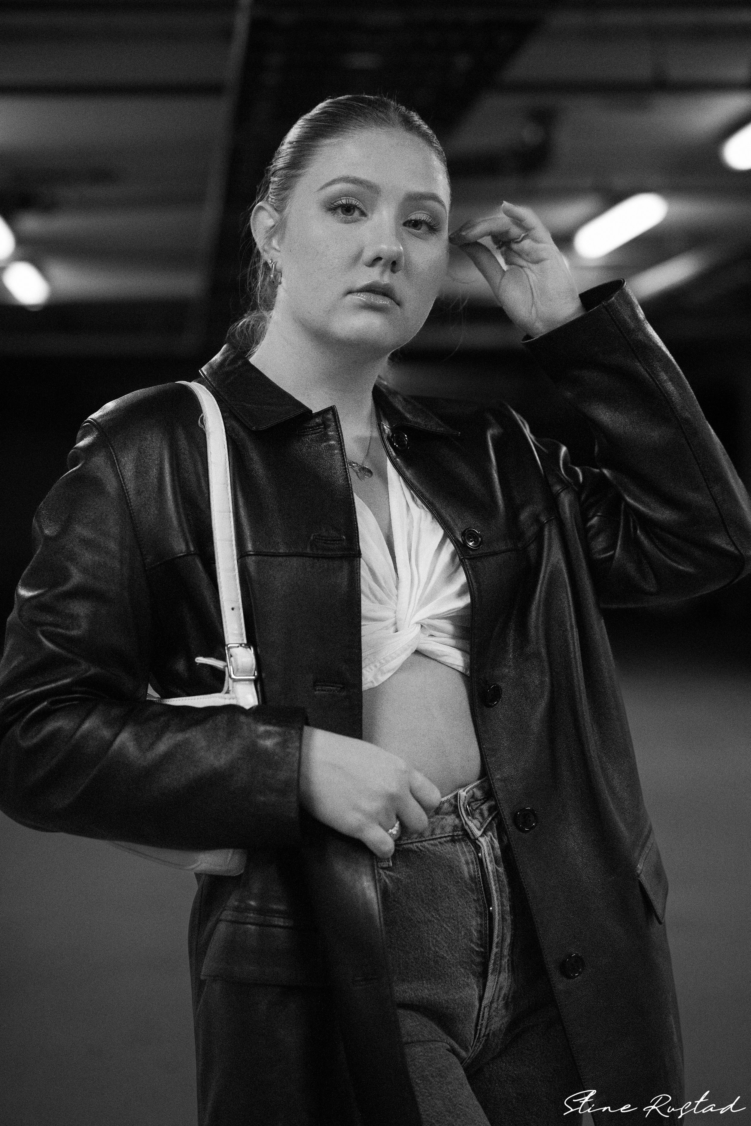 Black and white photo of a young woman with her hair pulled back, wearing a leather jacket, a knotted white top, and high-waisted jeans in an urban setting.