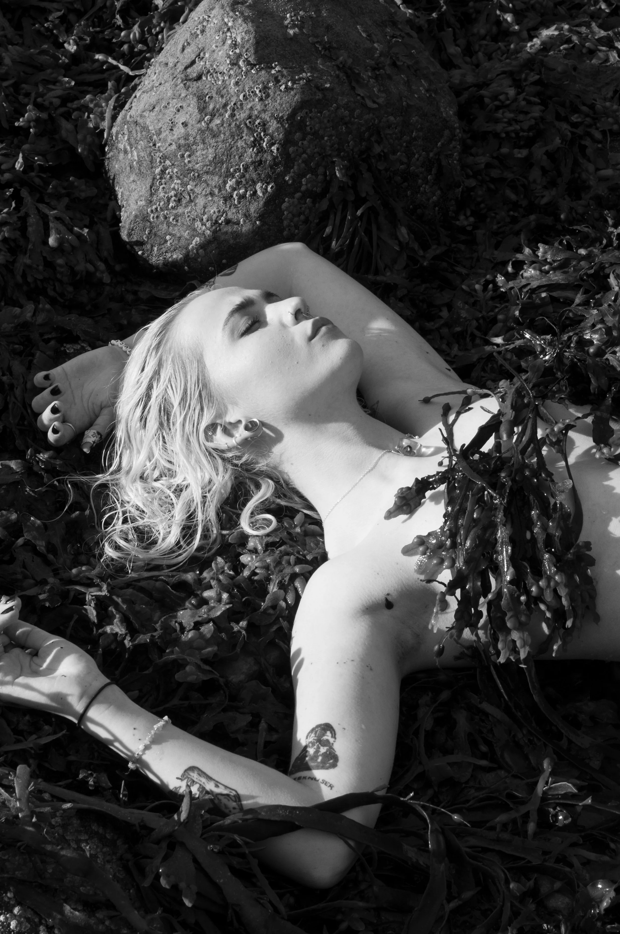 A woman lying on the ground among leaves and plants with a large rock nearby, her eyes closed, appearing to be relaxing or resting.