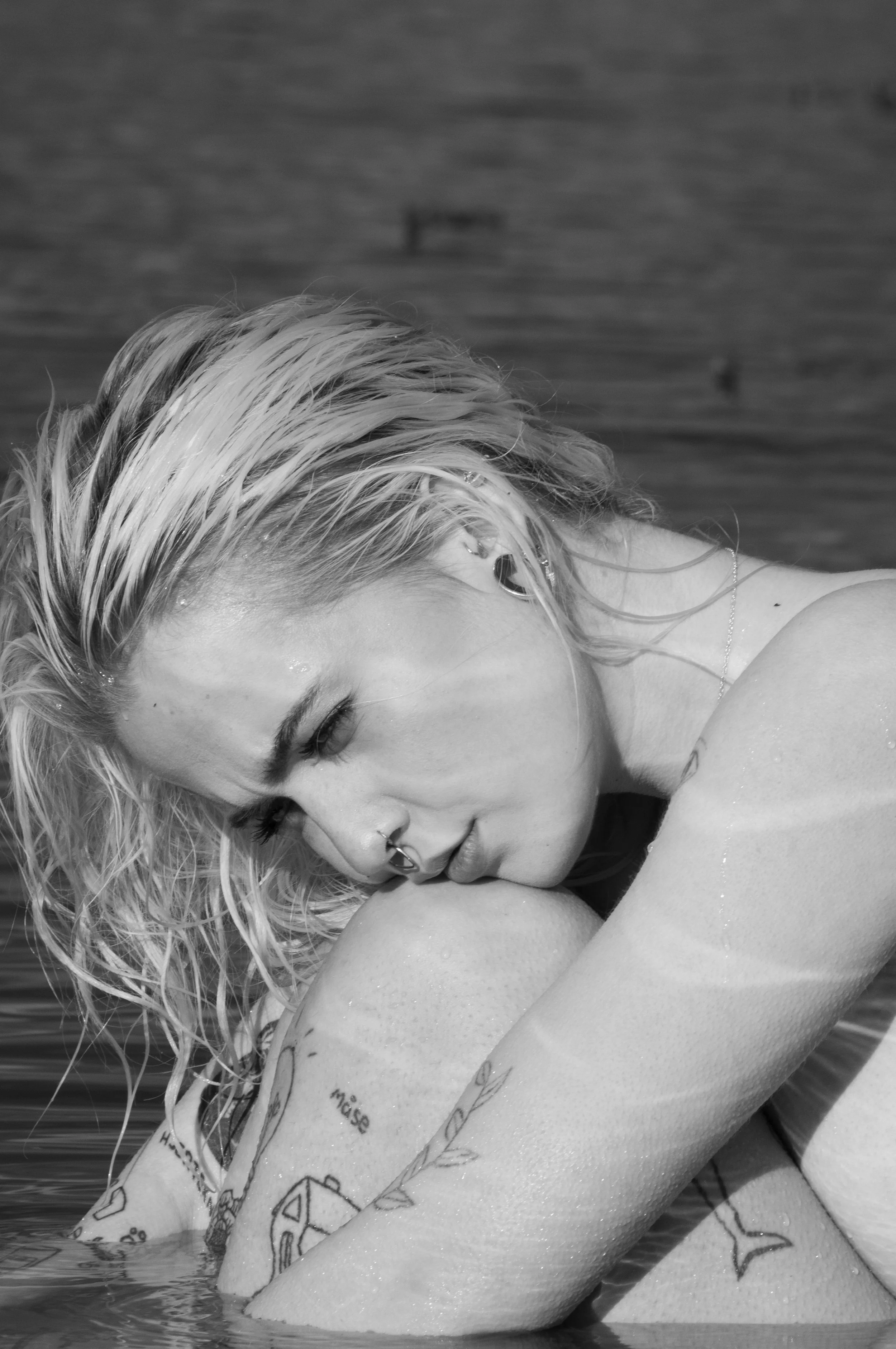 A black and white photo of a woman with wet blonde hair, hugging her knees while leaning on a body of water.