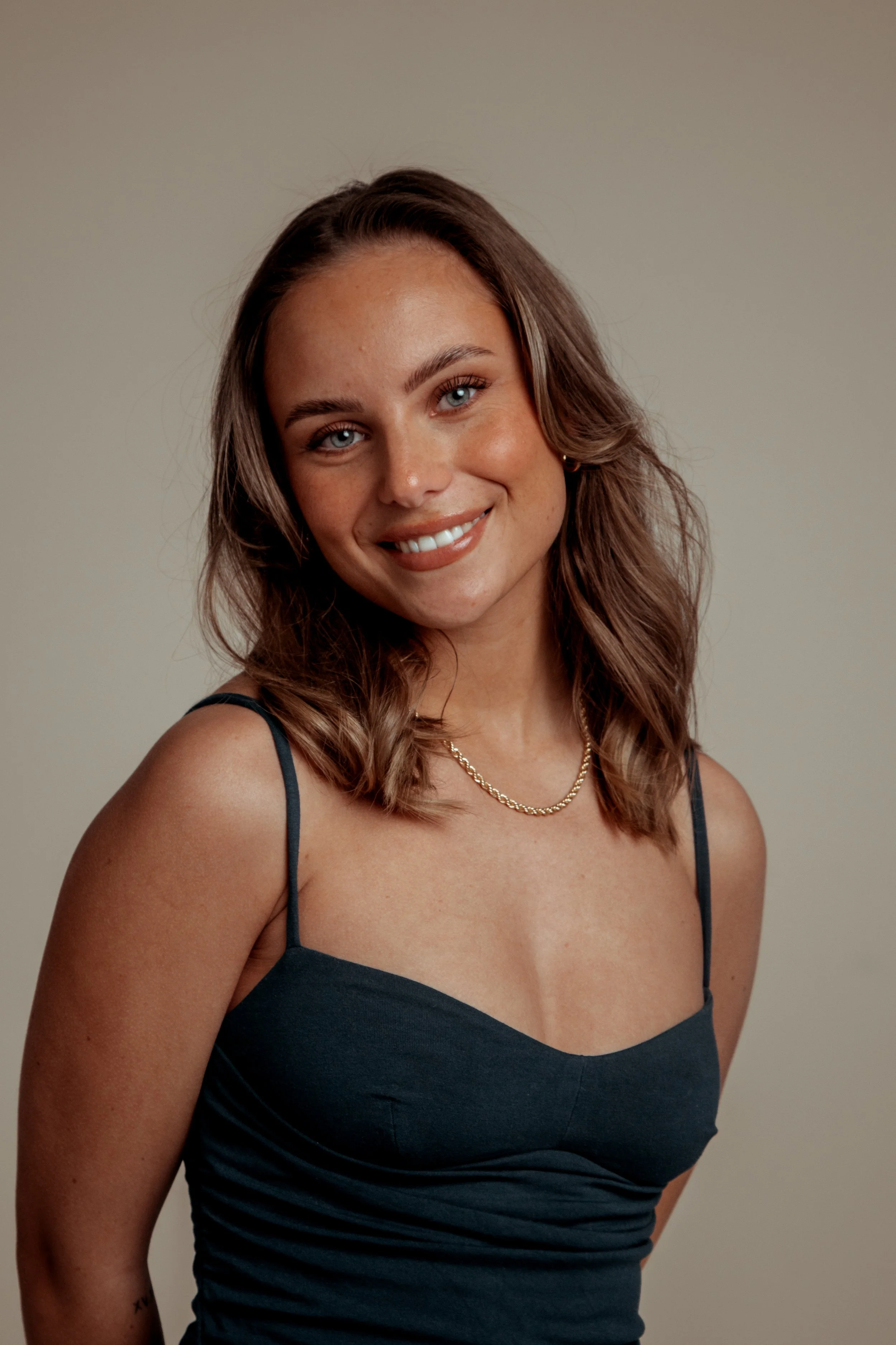 A young woman with shoulder-length wavy brown hair, blue eyes, and a warm smile, wearing a black spaghetti strap top and a gold chain necklace.
