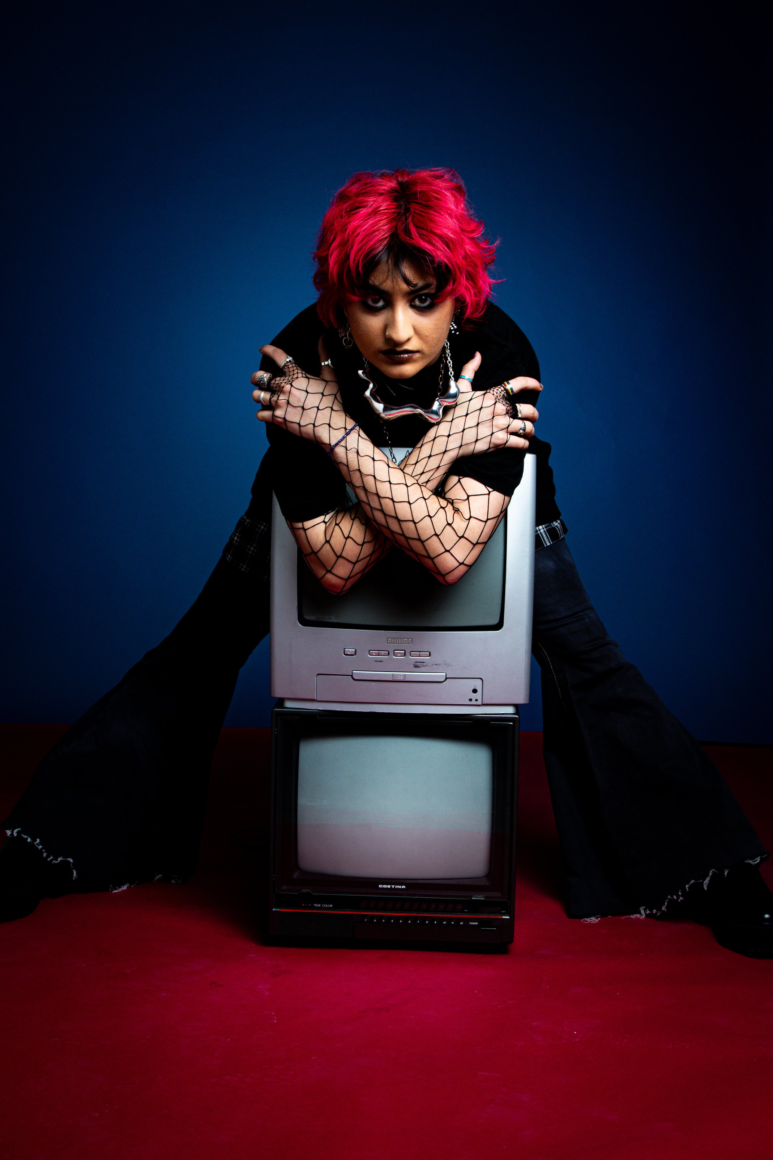 Person with bright pink hair leaning on CRT monitors, wearing black clothing and fishnet gloves, posing against dark blue background.