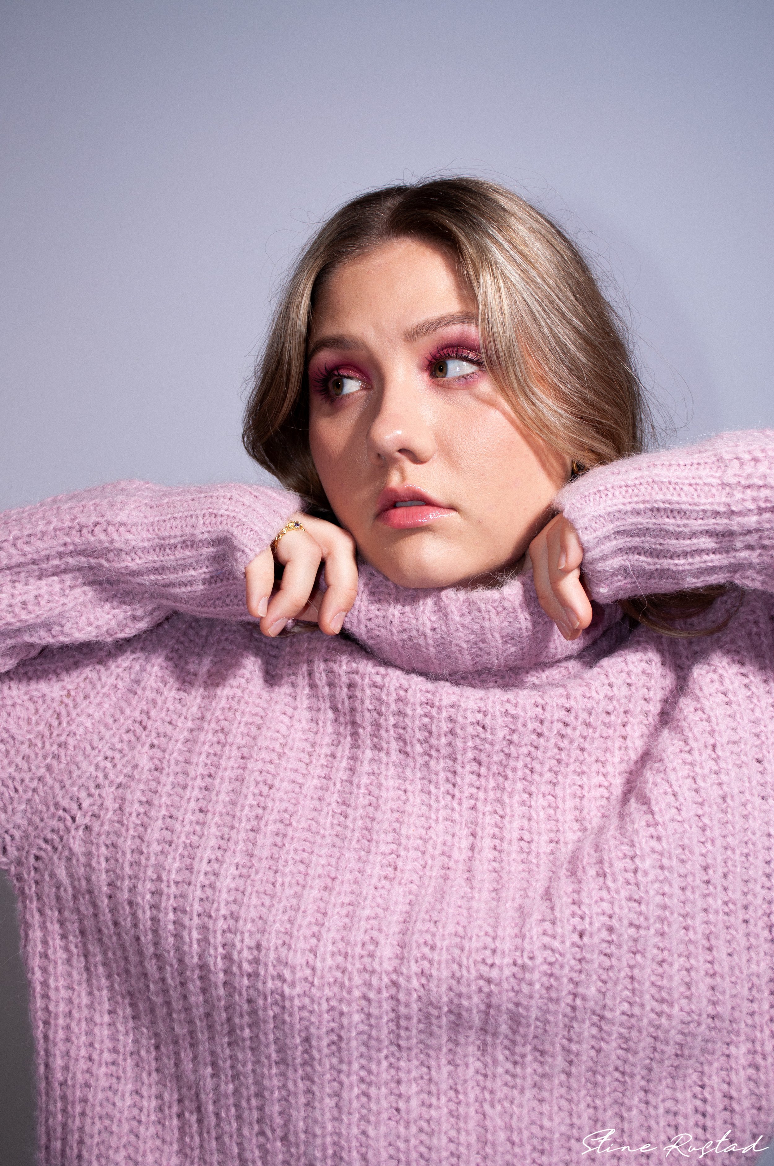 A young woman wearing a pink knitted sweater with her arms resting on her shoulders, looking to her left with pink eye makeup and wavy brown hair.