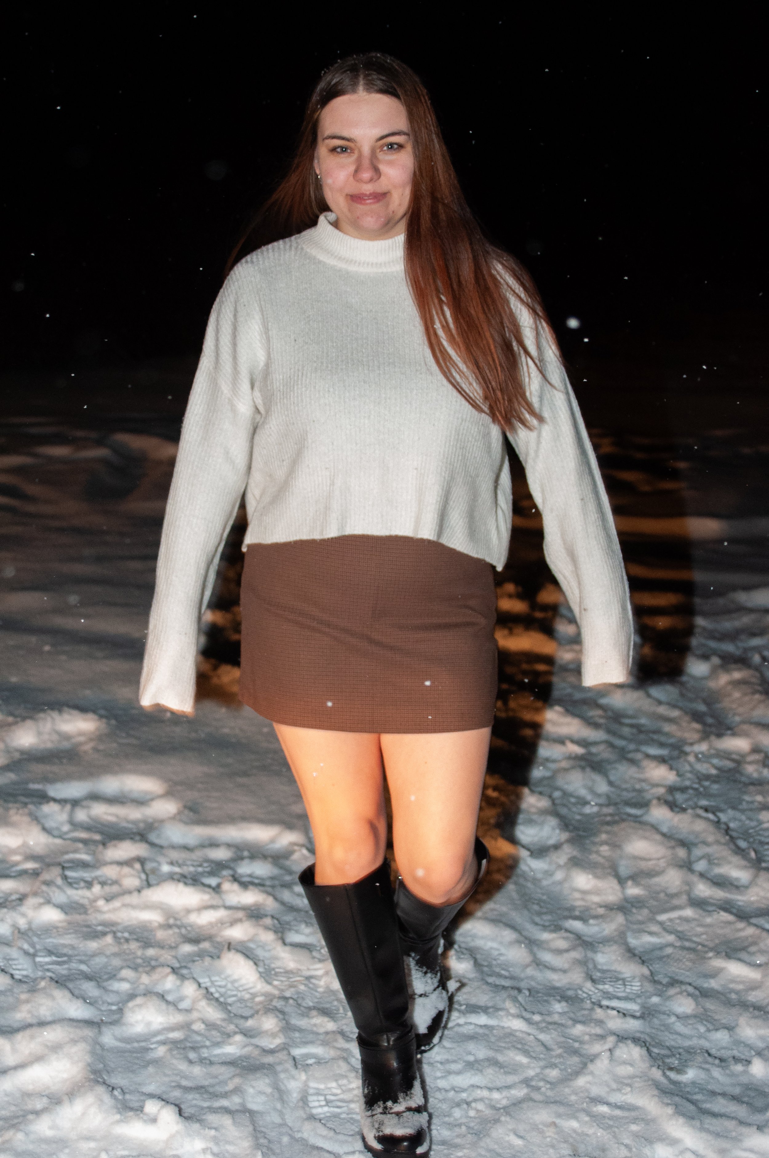 A woman with long red hair, wearing a cream sweater, brown skirt, and black boots, walking on snow at night.
