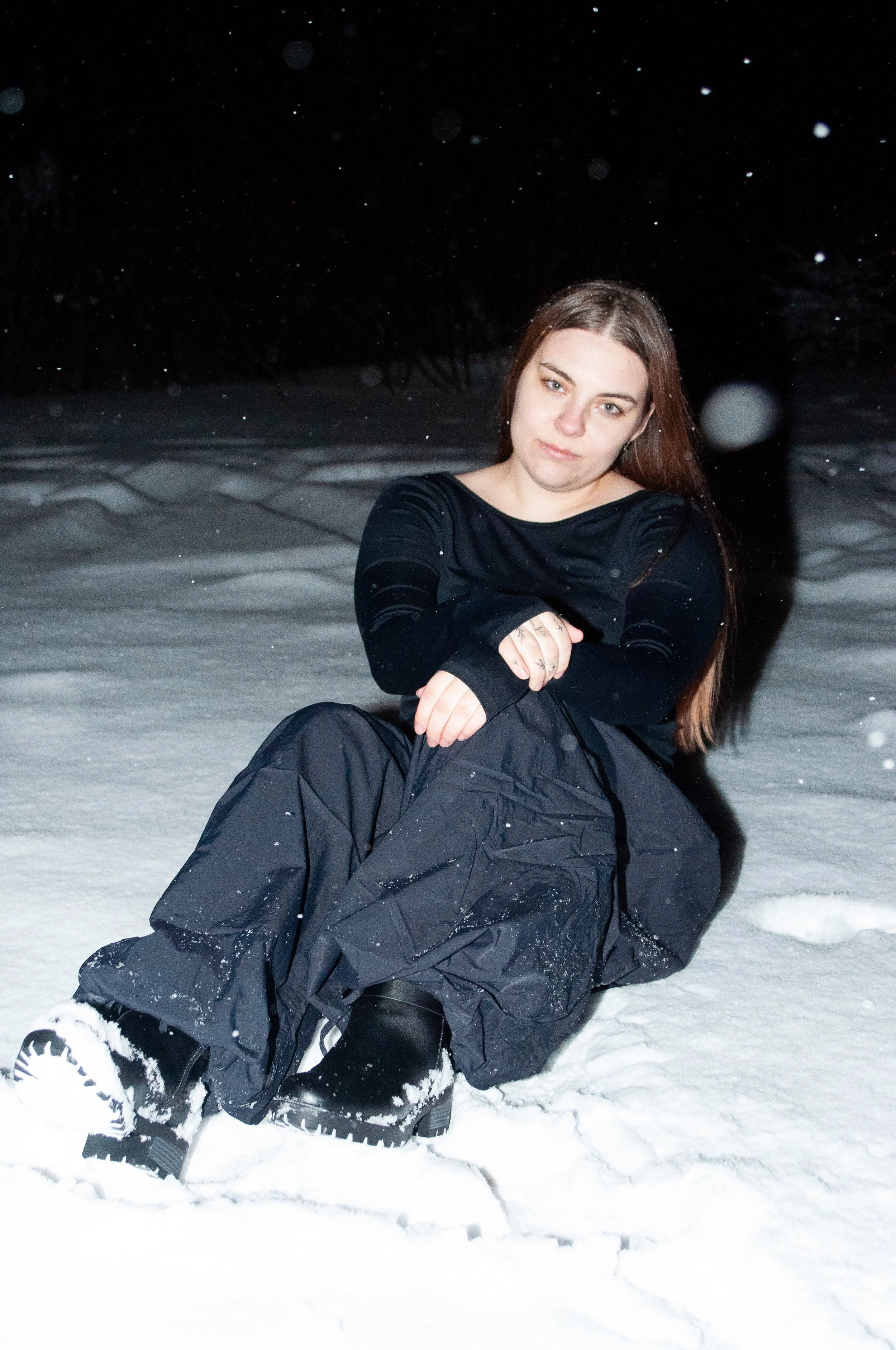 A young woman sitting in the snow at night, wearing black winter clothing and boots, with snow falling around her.