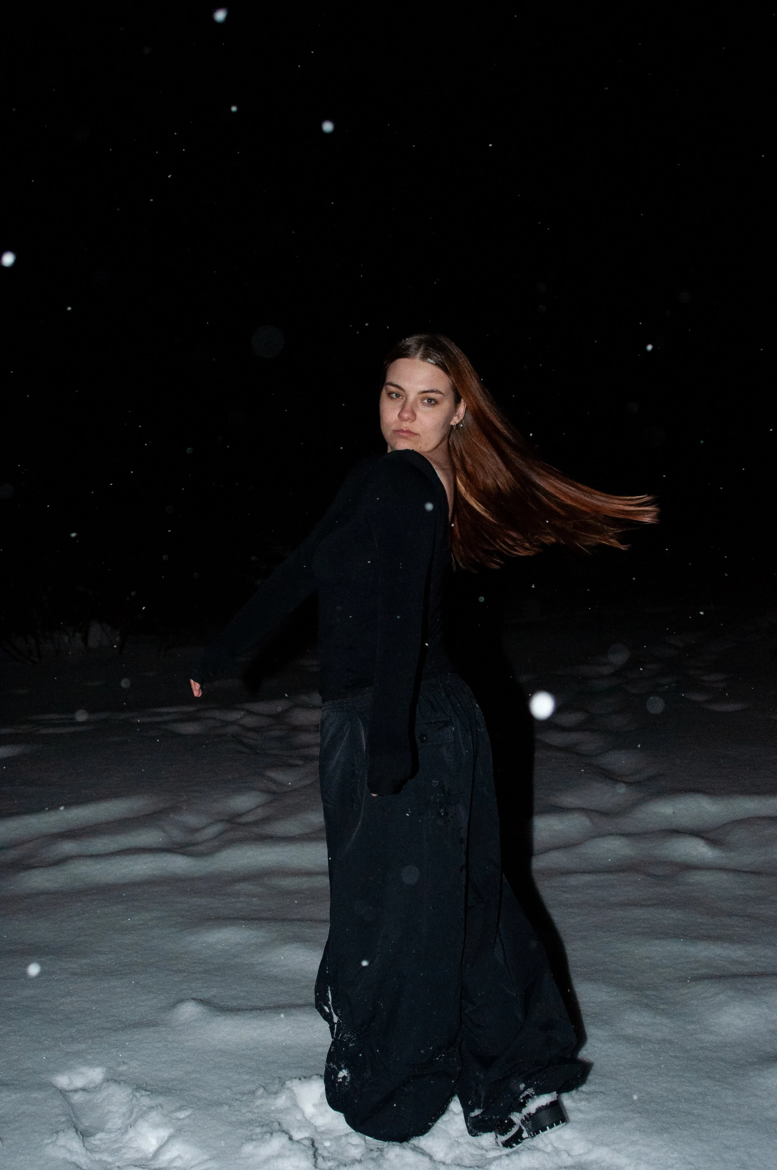 A young woman with long red hair standing in a snowy landscape at night, wearing black clothing.