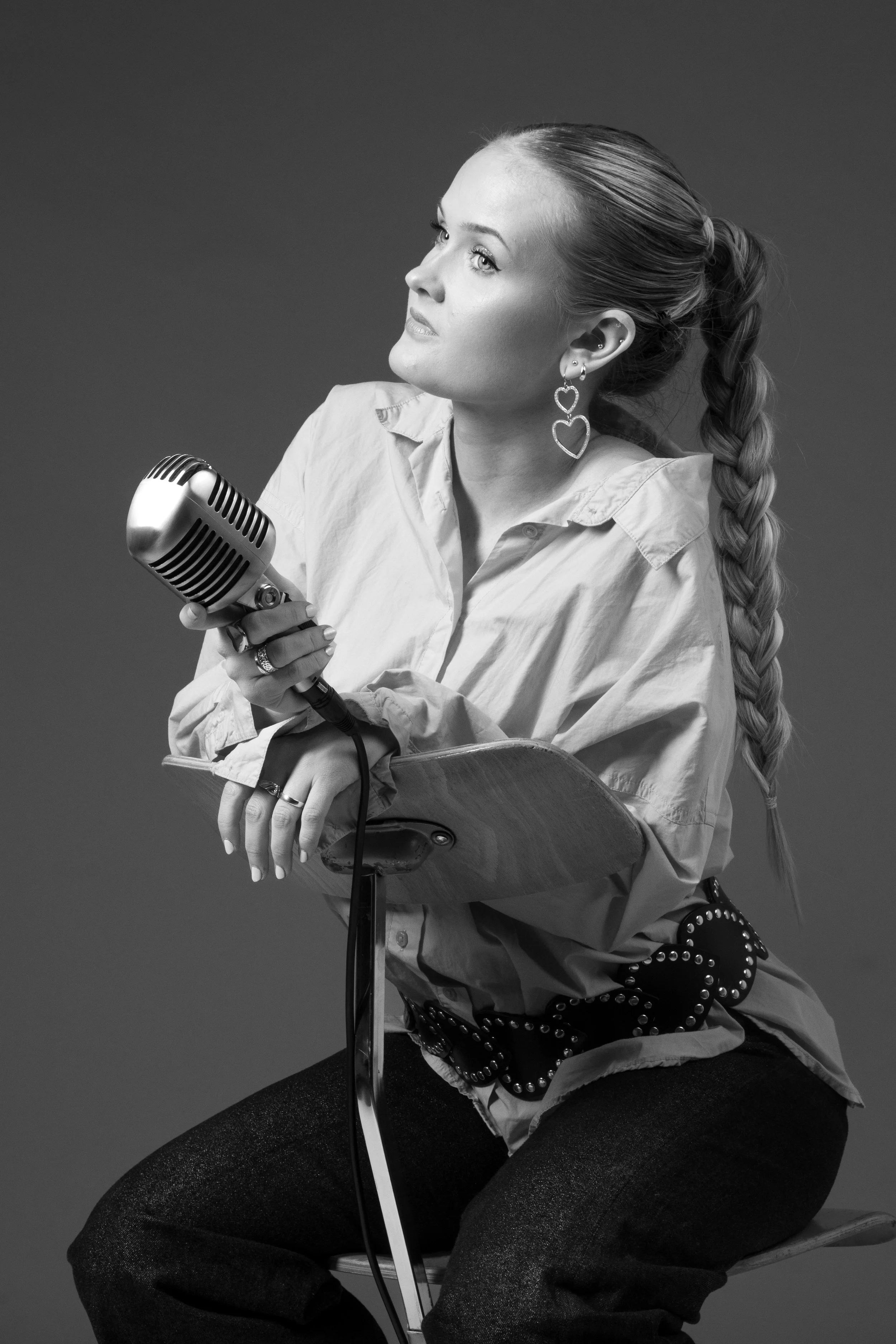 A woman with long braided hair, wearing a light-colored shirt and large heart-shaped earrings, holding a vintage microphone, sitting on a stool with a dark background.