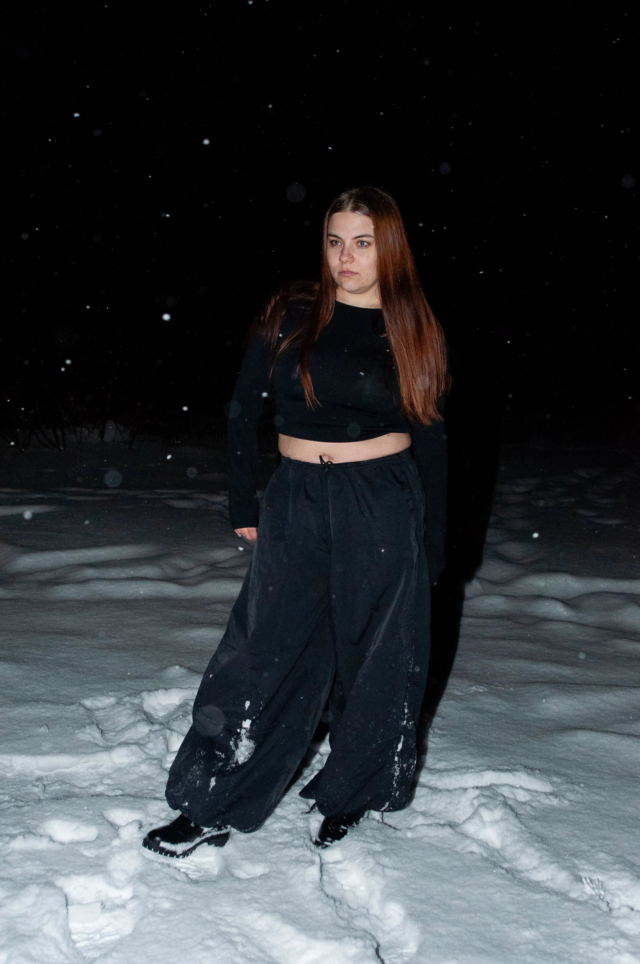 Young woman with long brown hair standing on snow at night, dressed in a black crop top and loose black pants.