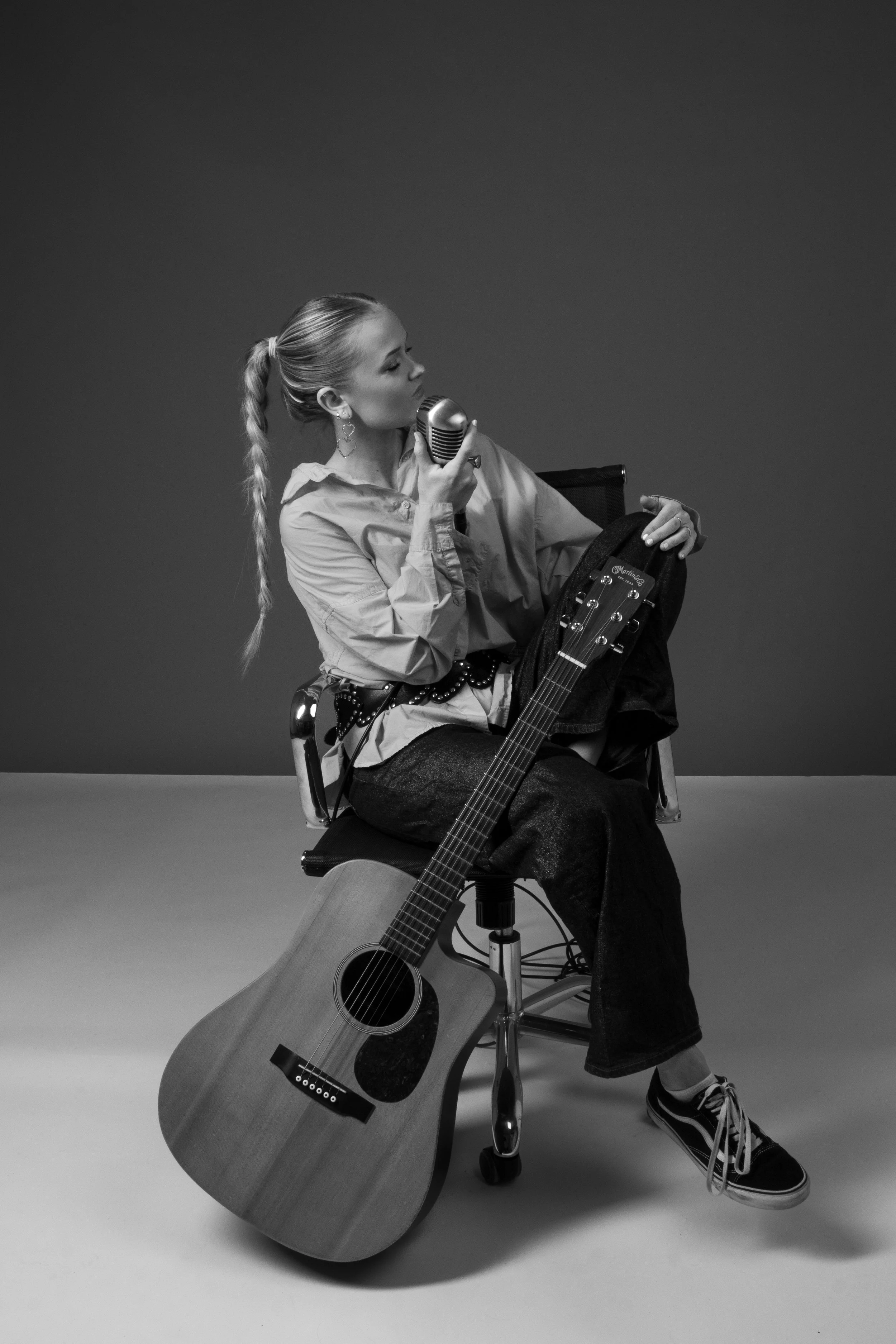 A woman with long braided hair sitting on a chair, holding a vintage microphone close to her lips, with an acoustic guitar resting on the floor in front of her, against a plain background.