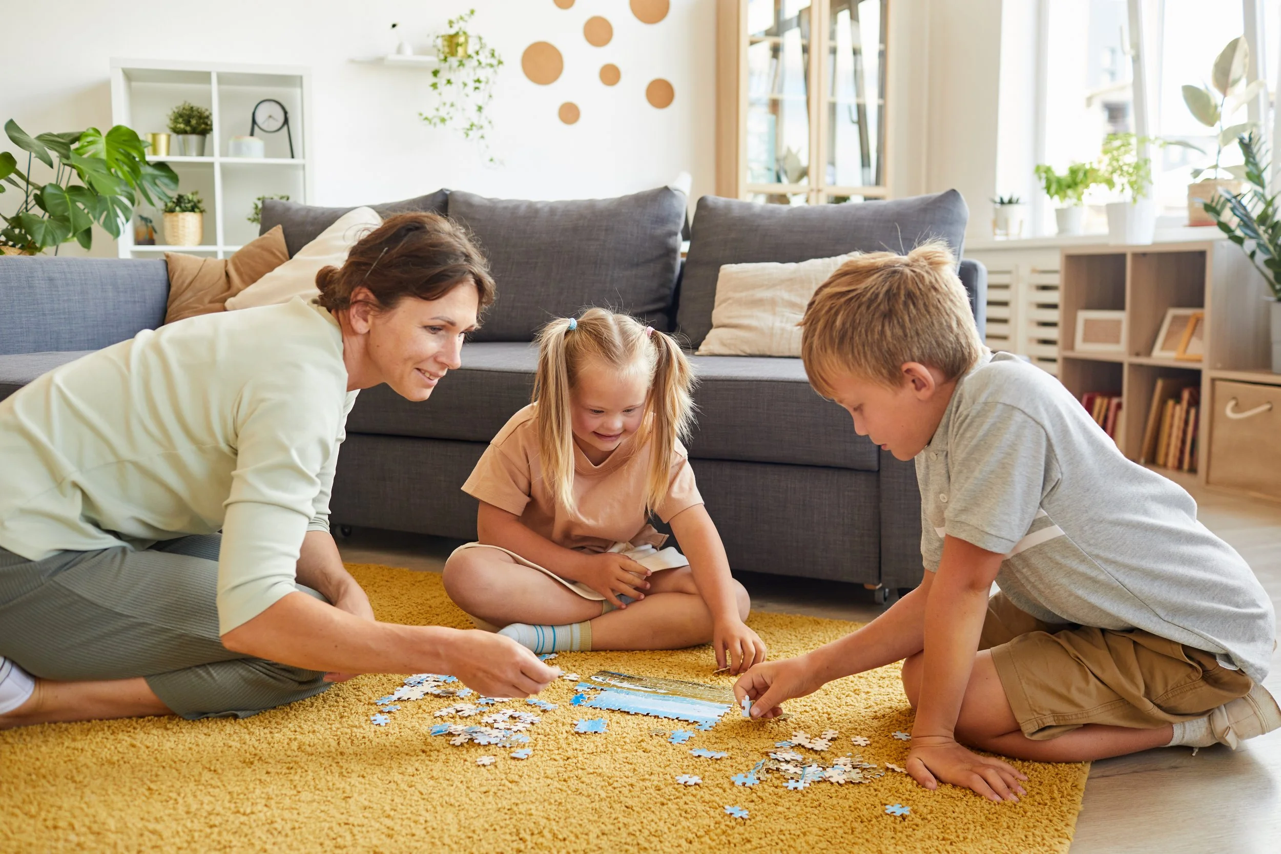 A woman, two children, and a boy sitting on a yellow carpet in a living room, working on a jigsaw puzzle together. The room has white walls, a gray sofa with beige and gray cushions, and various potted plants on shelves and windowsills.
