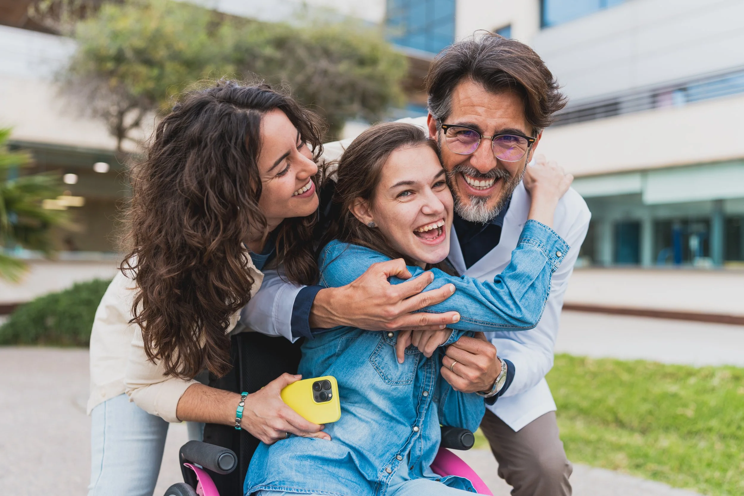 Four people, including a young girl in a wheelchair, smiling and hugging outdoors with modern buildings in the background.