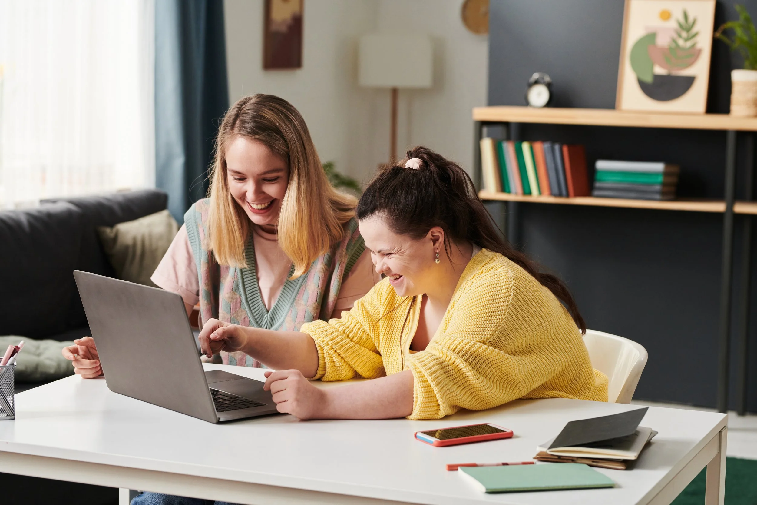 Two women smiling and laughing while looking at a laptop screen in a cozy home or office setting. They are sitting at a white table with notebooks, a smartphone, and pens, with a bookshelf and decor in the background.