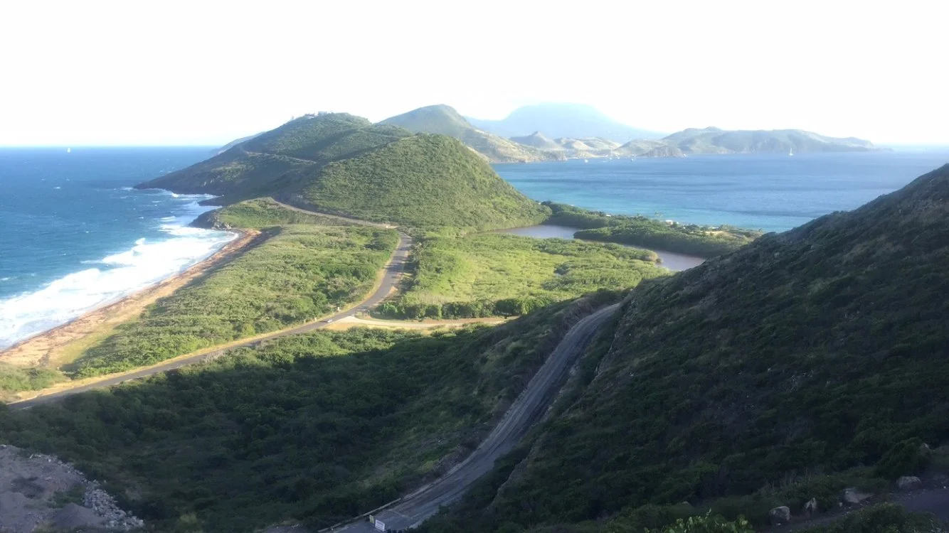 A scenic view of green mountains next to the ocean with winding roads and a harbor in the distance.
