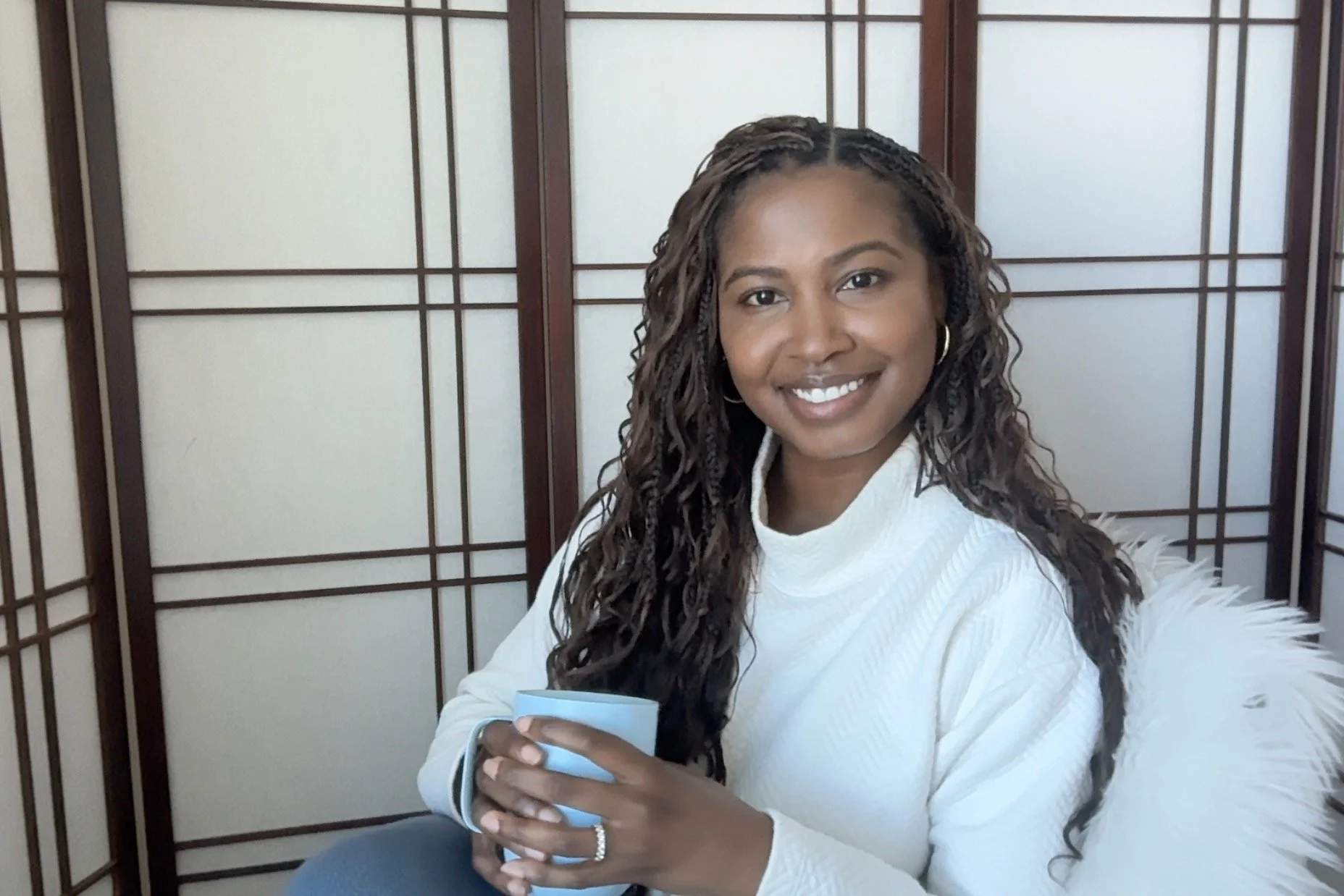 Picture of Dr. Pinard Morgan, a Black woman physician with long, curly hair smiling warmly and holding a light blue mug, sitting in front of a wall with wooden frames and frosted panels.