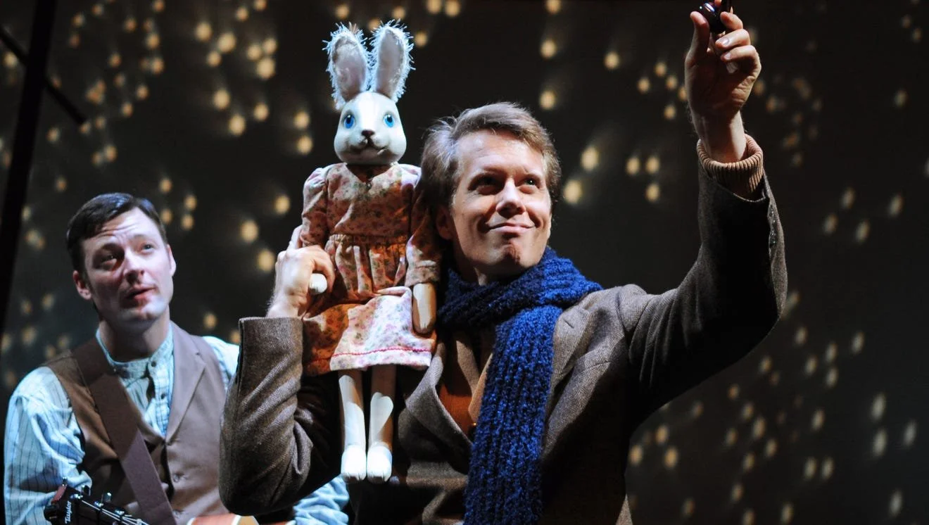 Two men on stage, one with a toy rabbit on his shoulder, performing in a theatre with sparkling lights in the background.