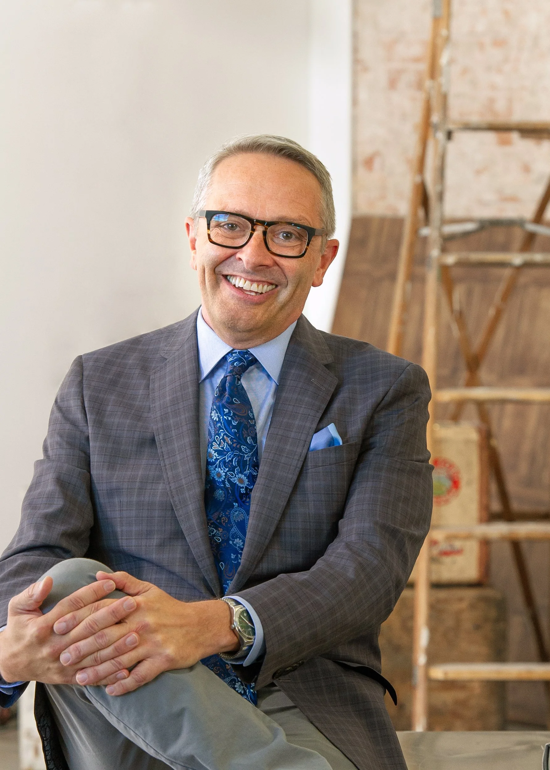 Dwayne Hartford wearing glasses, a gray plaid blazer, a blue dress shirt, and a patterned blue tie, sitting with his hands clasped in front of him in a room with an unfinished brick wall and wooden scaffolding in the background.