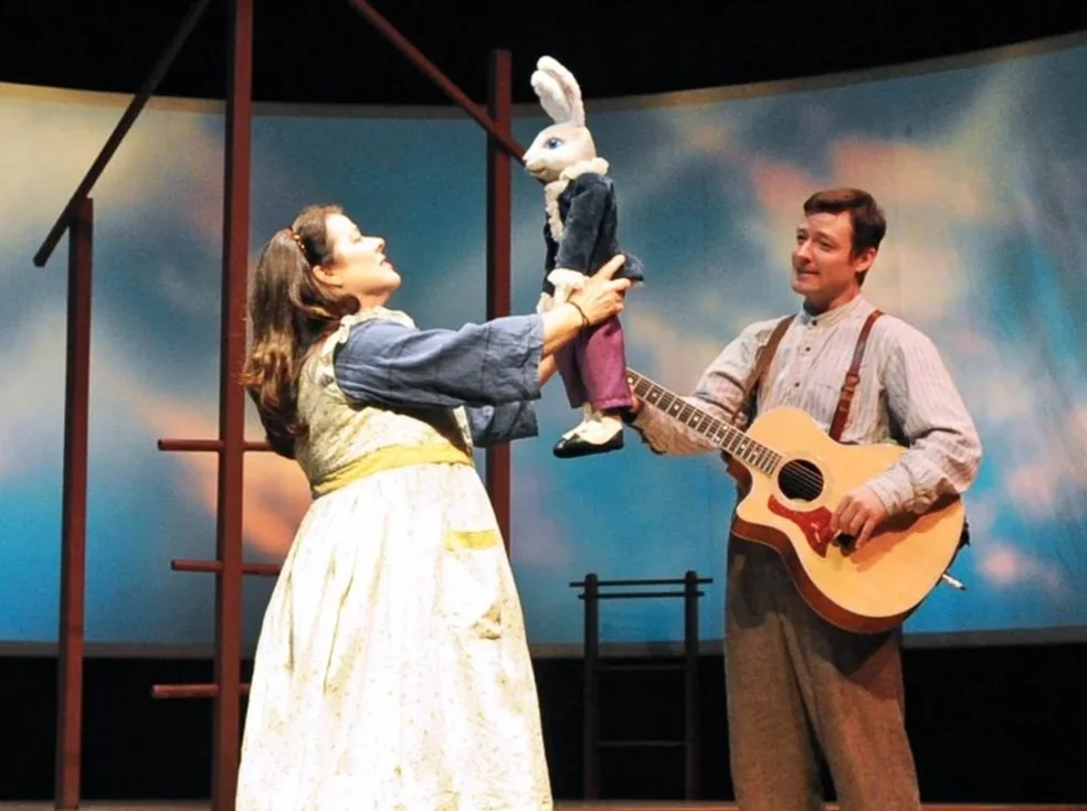 Two performers on stage, one woman holding a toy rabbit and a man playing an acoustic guitar. The background features a painted sky with clouds.