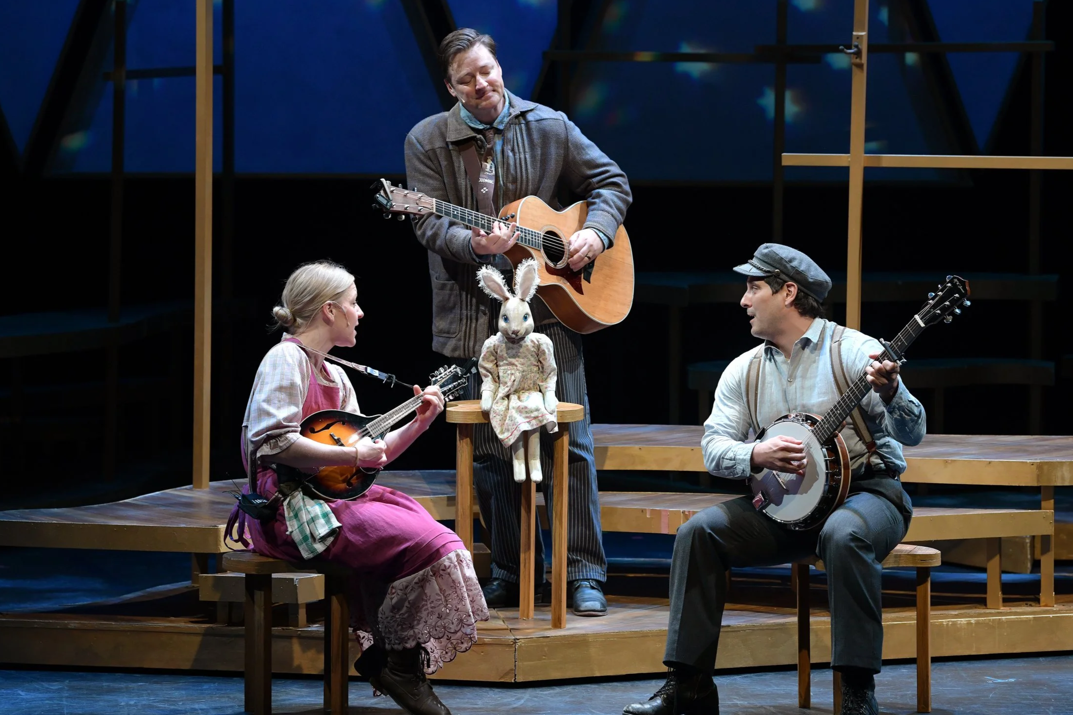 Two performers on stage, one woman holding a toy rabbit and a man playing an acoustic guitar. The background features a painted sky with clouds.