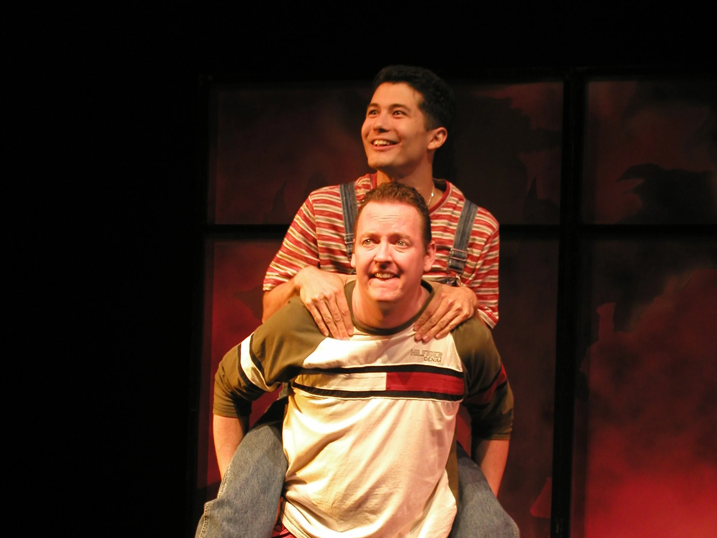 Two men performing in a play, one sitting on the other's shoulders. The man on top is smiling and wearing a striped shirt, while the man below has a more serious expression and is wearing a color-blocked shirt.