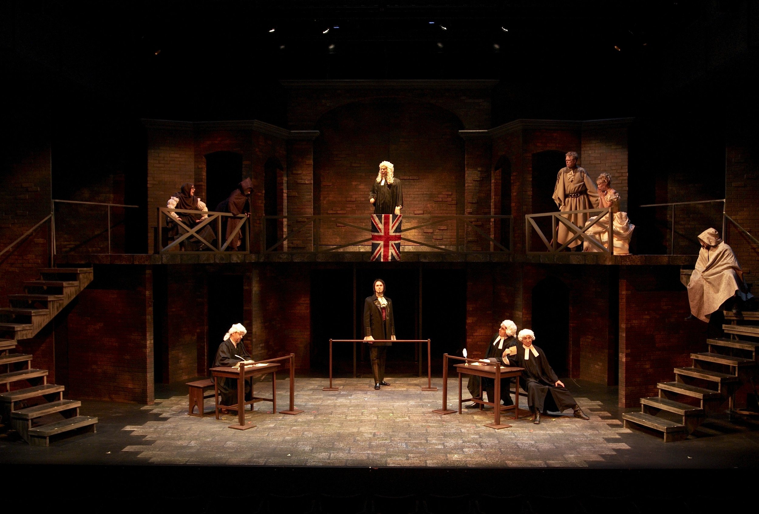 A theatrical stage setup resembling a courtroom with actors dressed in period costumes, including judges, lawyers, and officials, with a backdrop of brick walls and stairs on both sides.