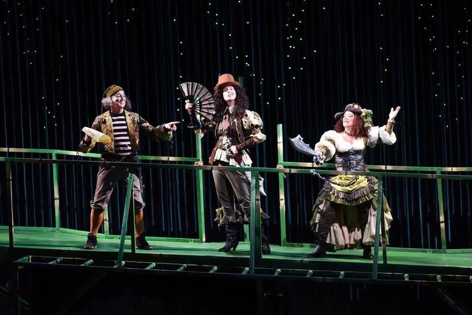 Three performers dressed in colorful, vintage costumes on a theatrical stage with a dark background and string lights, engaging in a theatrical performance.