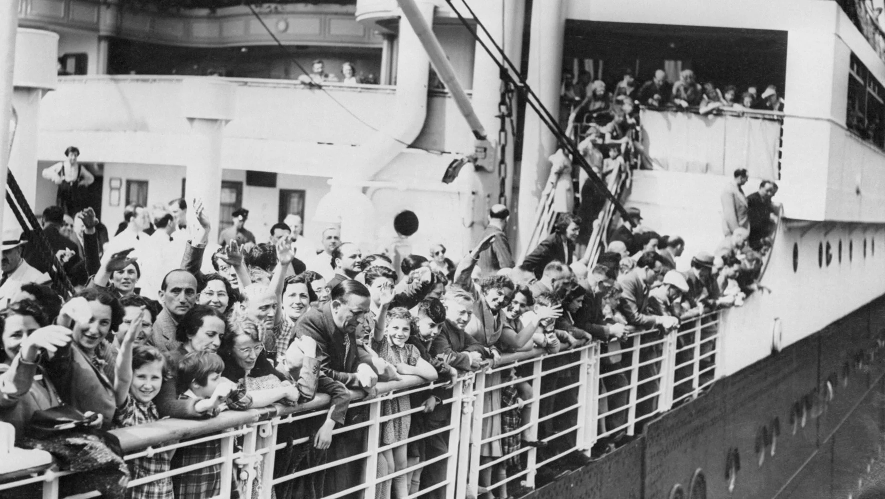 Black and white photo of a crowd of people on the deck of a ship, leaning on the railing and waving. Some are smiling and looking at the camera. The ship has multiple levels with passengers visible on the upper decks.