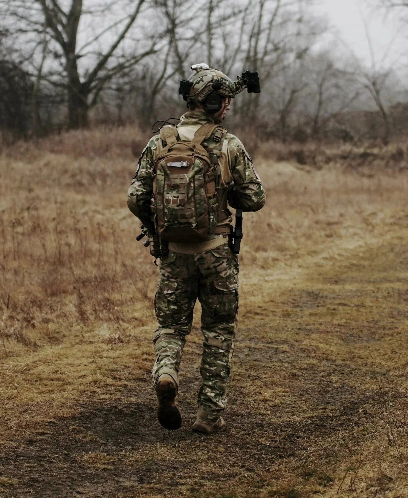 A soldier dressed in camouflage uniform and helmet, walking through a field with leafless trees in the background.