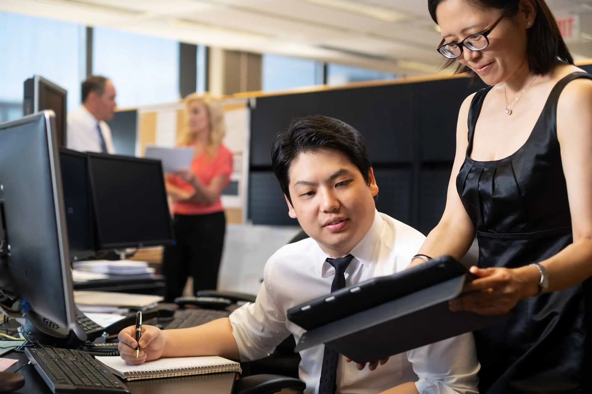A man in business attire sitting at a desk, looking at a tablet held by a woman in a black dress, in an office with computer monitors and colleagues working in the background.