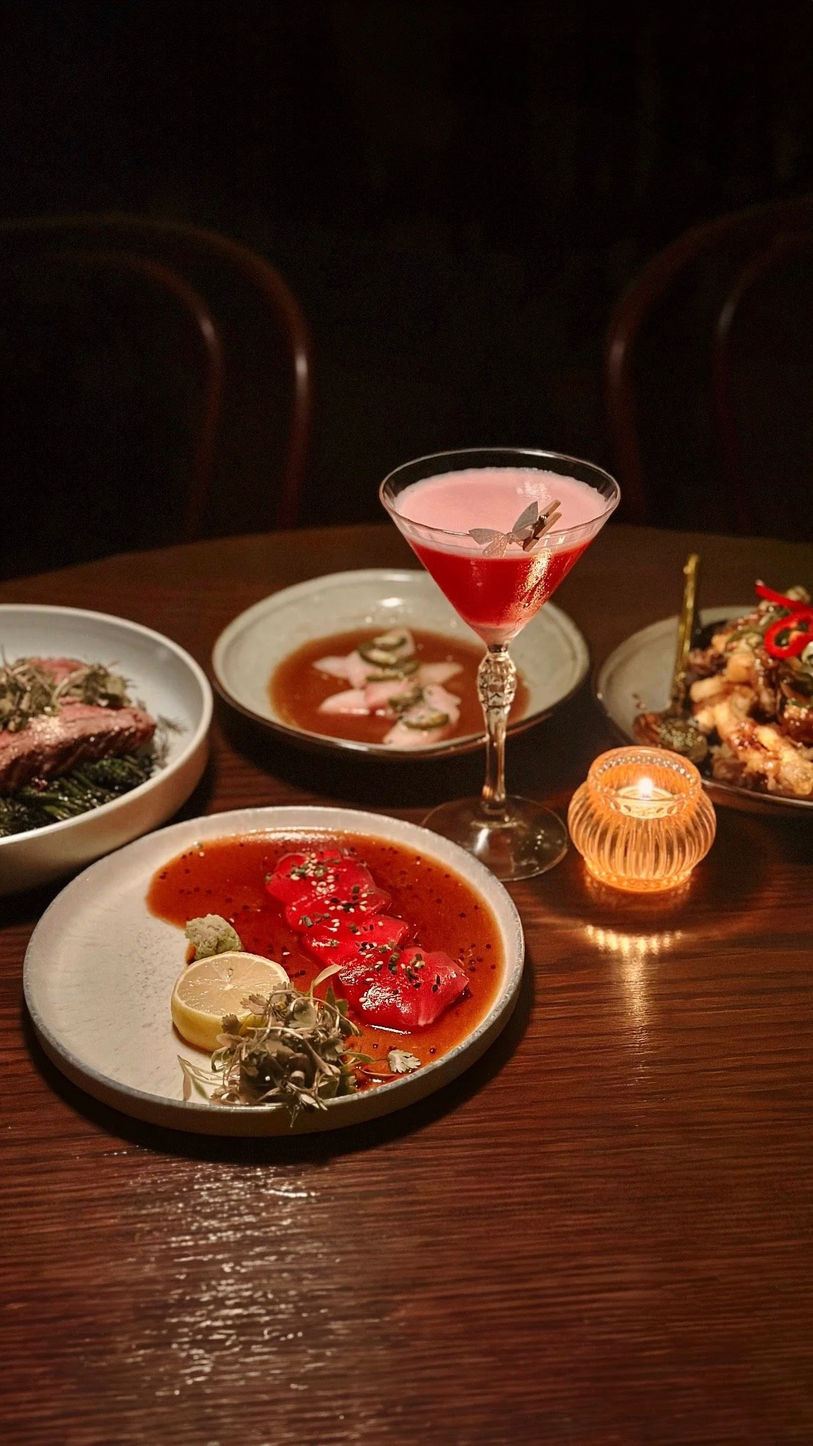 Several plates of sashimi and a fuchsia cocktail drink on a dark wood table.