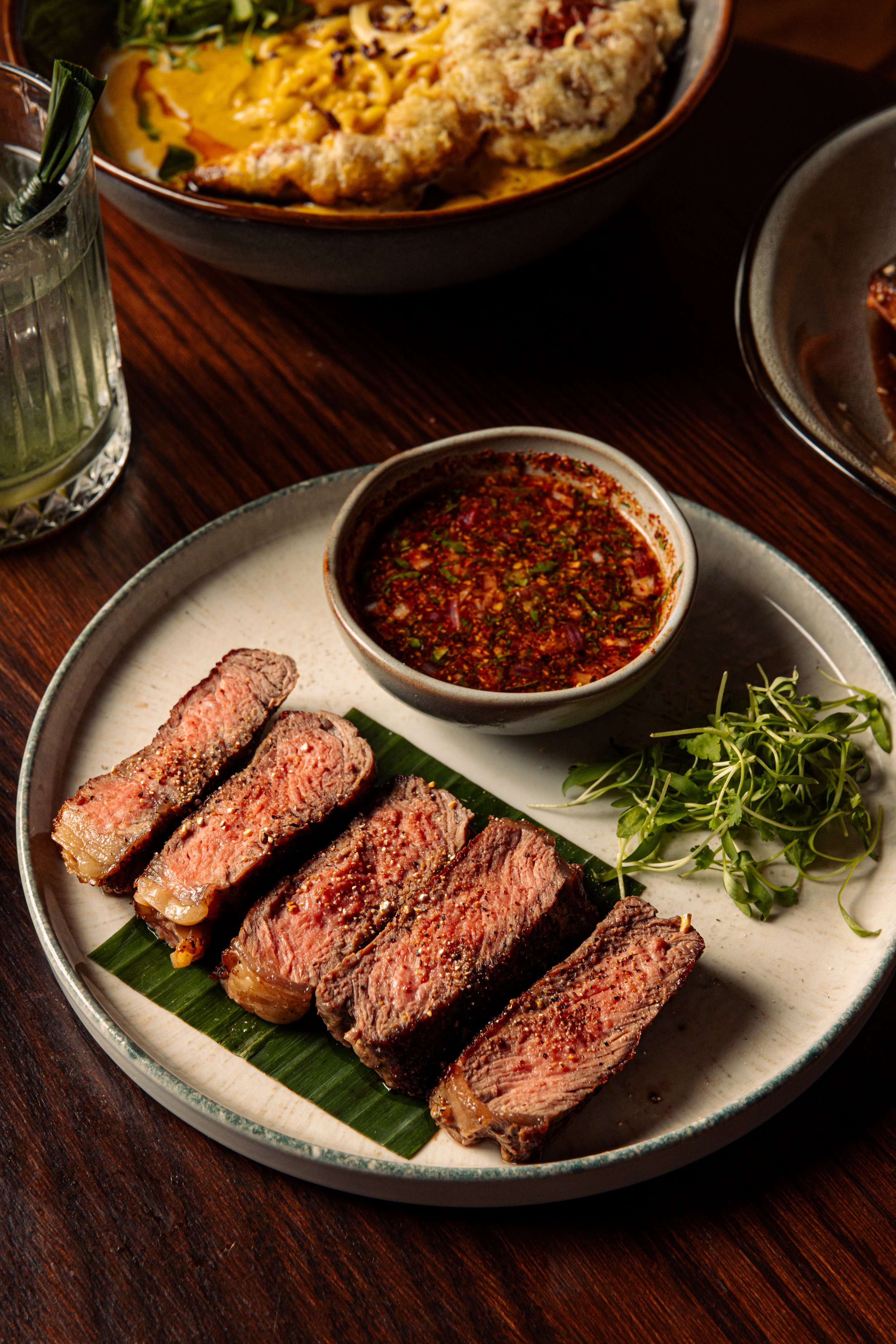 Plate of medium rare steak slices, micro cilantro, and a bowl of spicy, chili sauce.