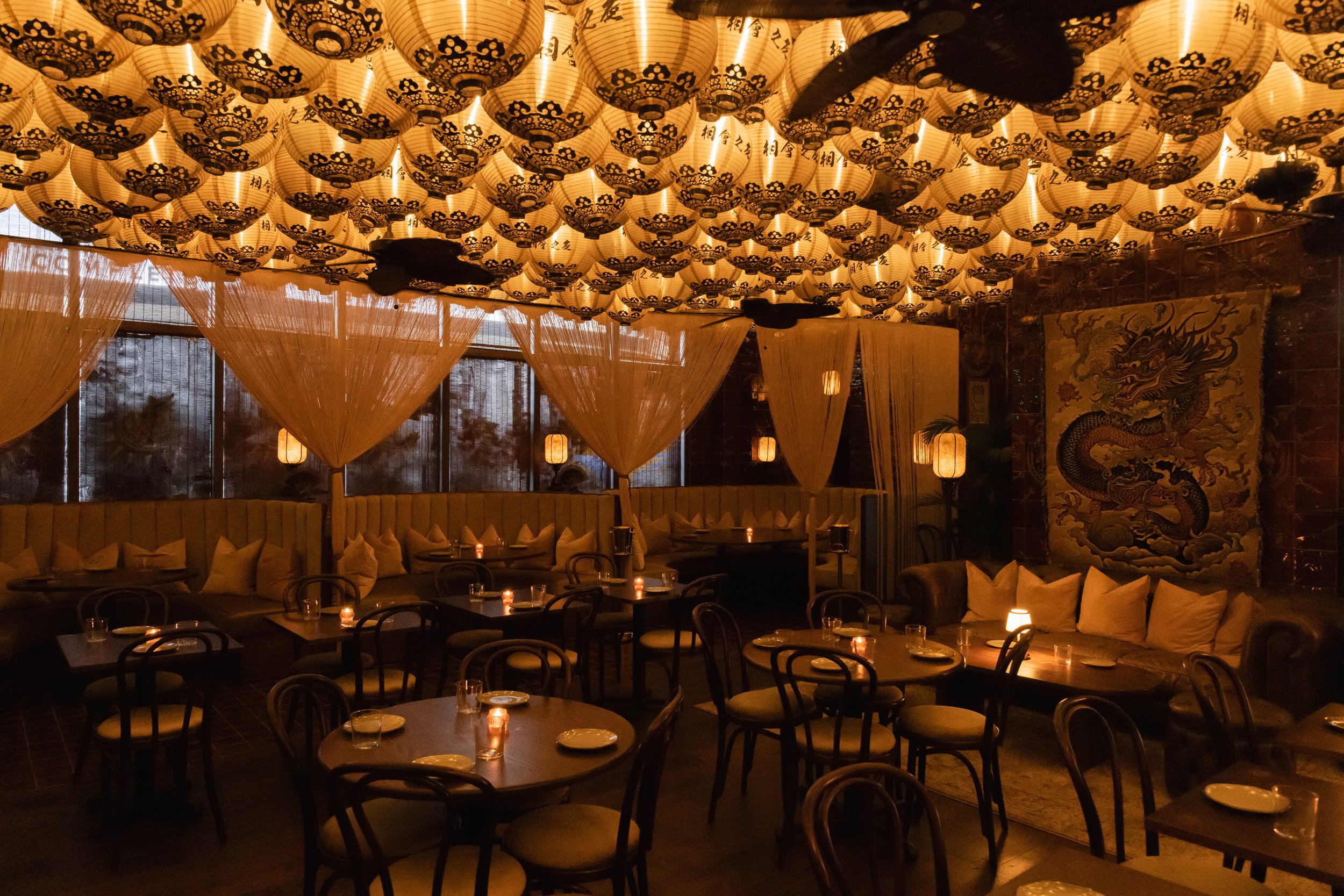 Curtained booths and wooden tables in The Night We Met’s dining room. A tapestry of a dragon hangs on the wall above a leather couch.