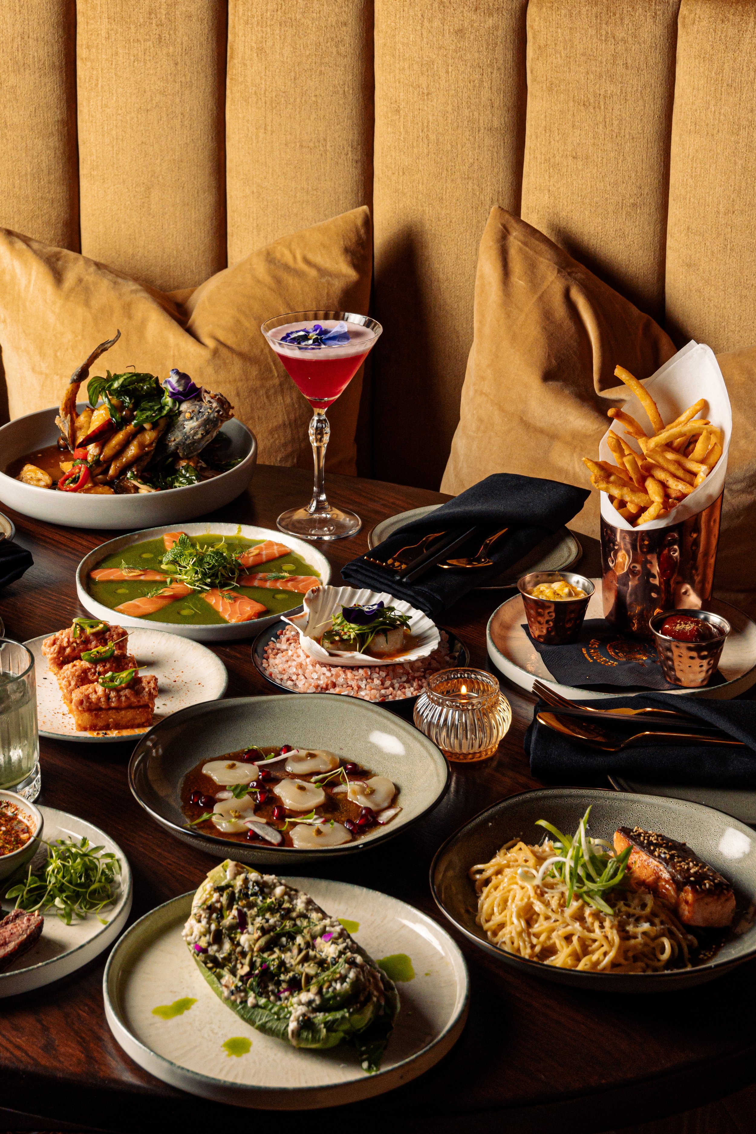Table set with cocktail drinks and various Pan-Asian dishes: salmon crudo, crispy rice, garlic noodles, and branzino.