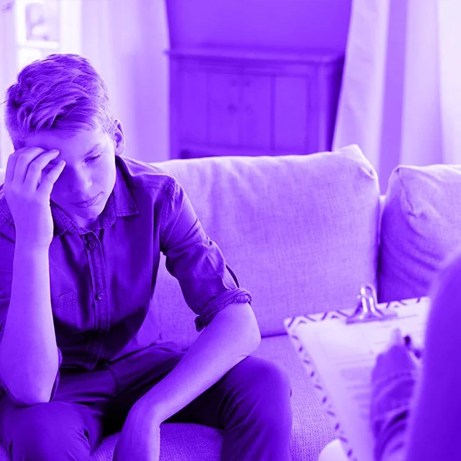 A young boy sitting on a couch, looking worried or anxious, with a person holding a clipboard in front of him, in a room with purple lighting.