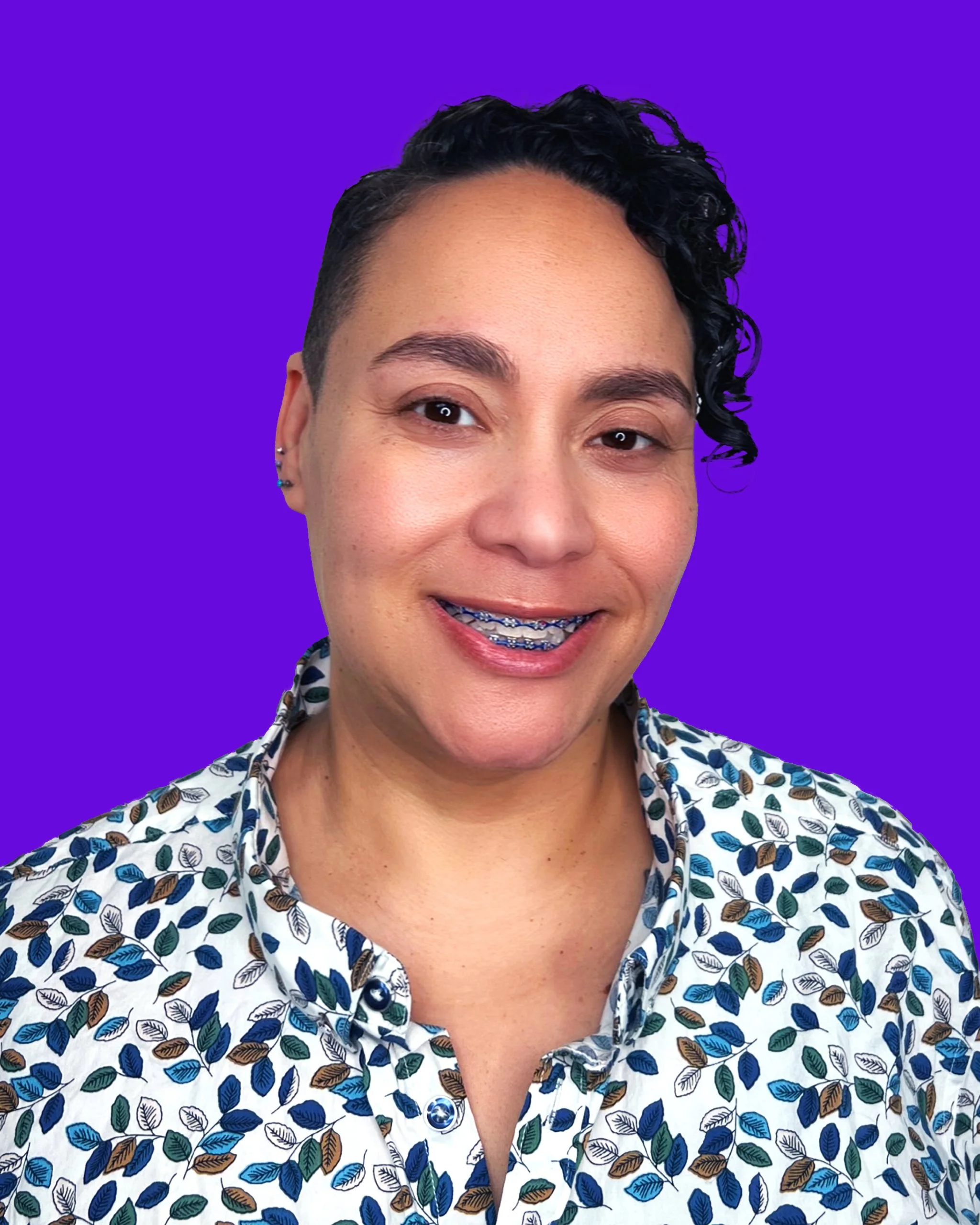 Close-up of a woman with short curly black hair, wearing braces, smiling and dressed in a patterned shirt with blue, green, and brown leaves on a white background, against a vibrant purple background.