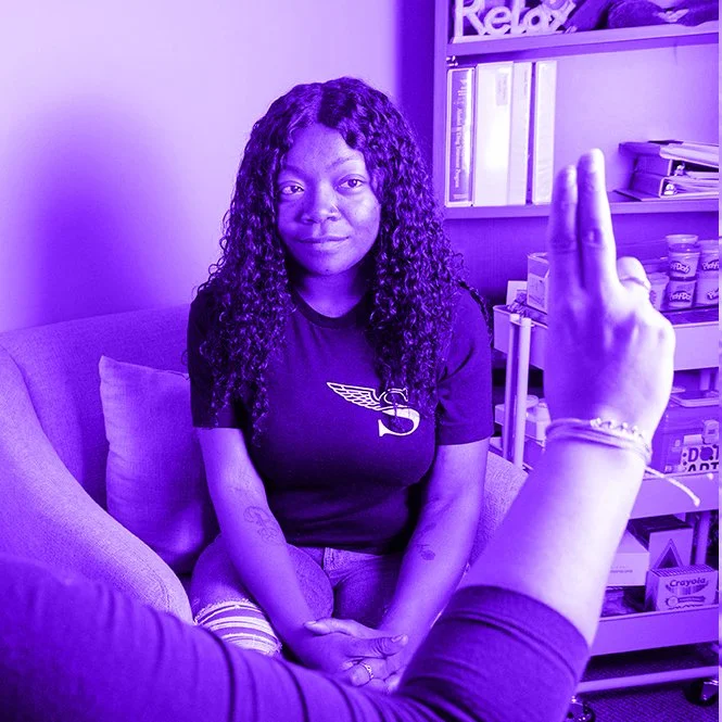 A woman sitting on a couch with long curly hair, wearing a black T-shirt, and a person in the foreground holding up two fingers in a peace sign, in a room with books and shelves.