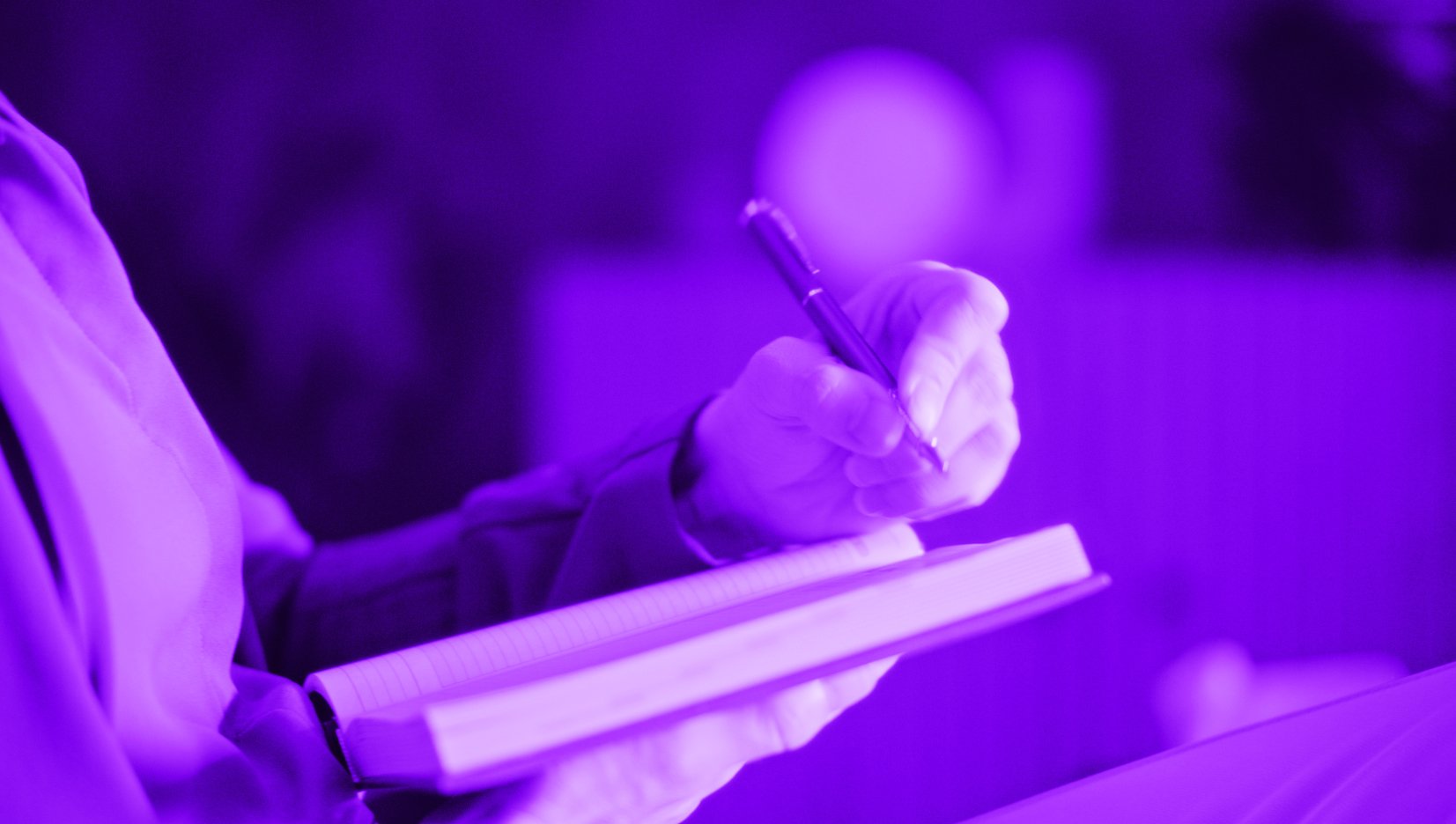 A person wearing gloves writes in a notebook with a pen, illuminated by purple lighting.