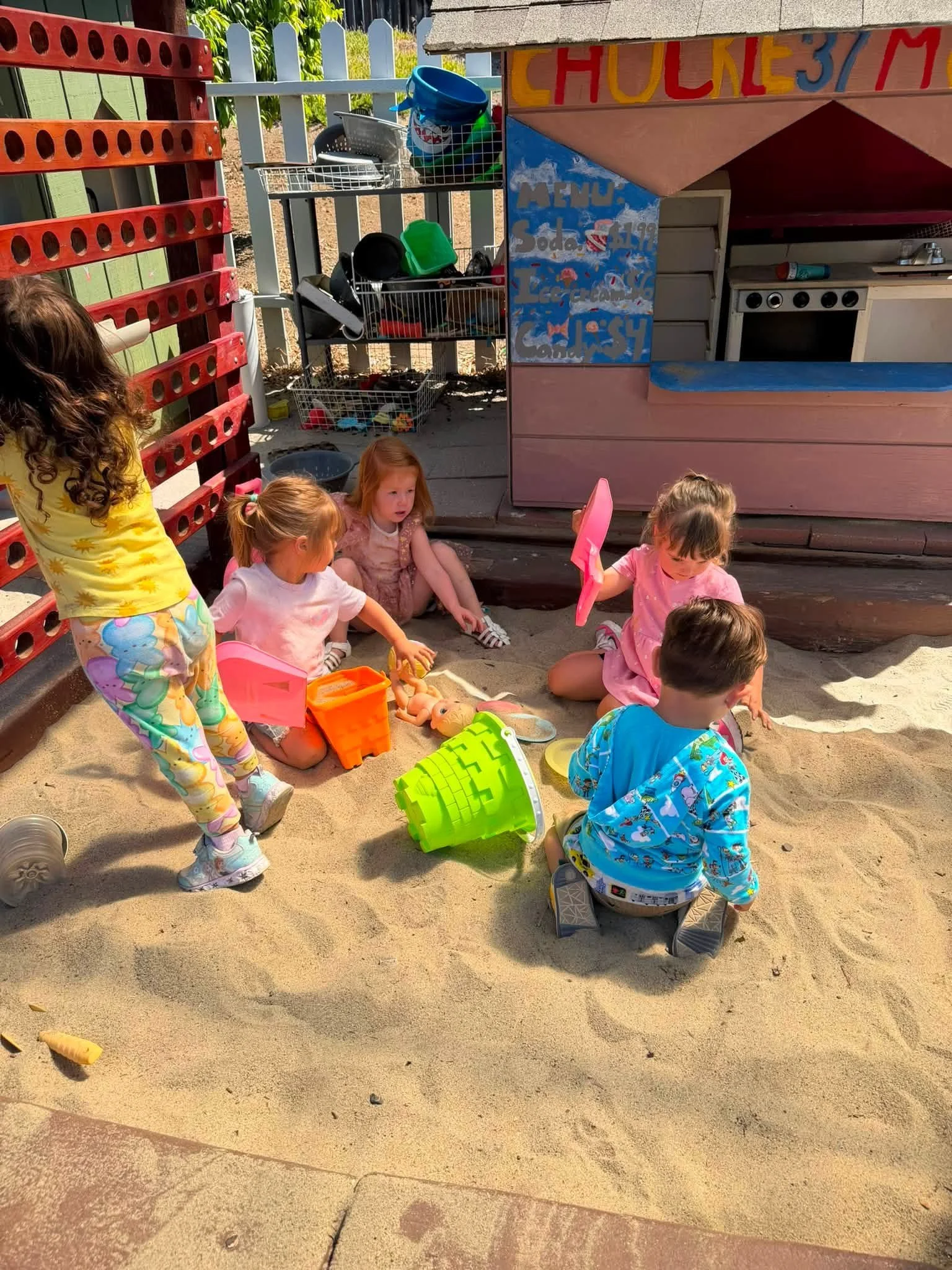 Children playing in a sandbox at a playground with toys, at a puppet stand labeled 'Chucky's.'