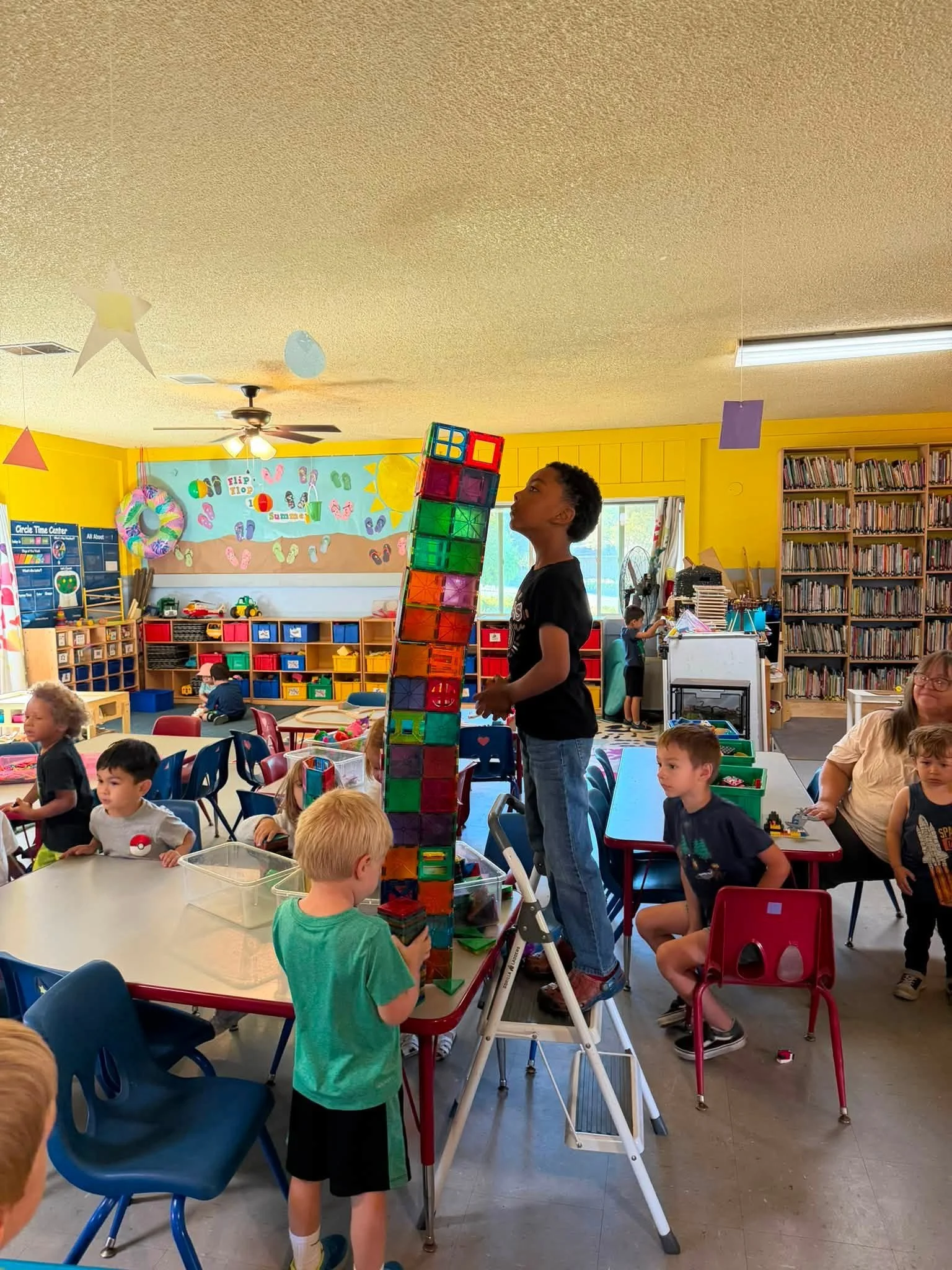 Children playing with a large tower of colorful building blocks in a classroom decorated with sea-themed artwork and bookshelves.