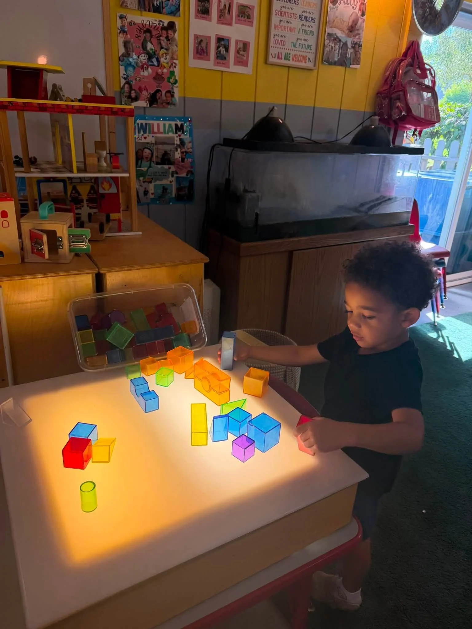 A young boy with curly hair playing with colorful translucent building blocks on a lit-up table in a classroom or playroom. Background includes bright yellow and pink wall decorations and educational posters, with a pink backpack hanging near a window.