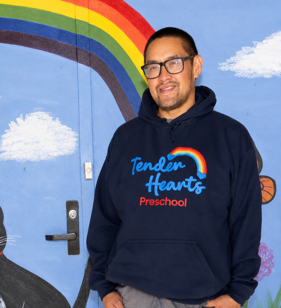 A teacher smiling standing in front of a colorful mural with a rainbow, clouds, flowers, a butterfly, a cat, and a ladybug.