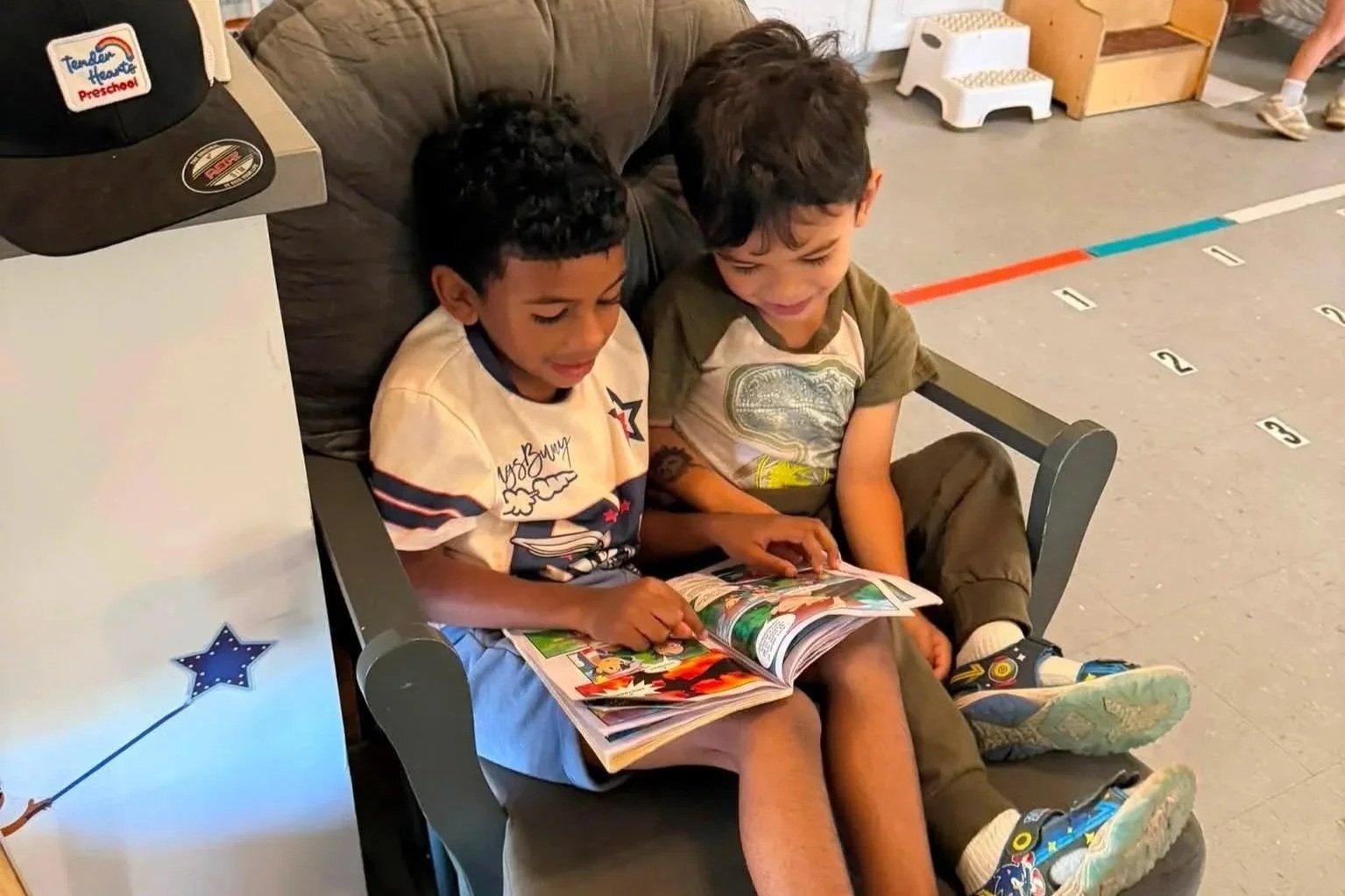Two young boys sitting closely on a black chair, sharing and reading a colorful comic book, smiling and enjoying their time together.