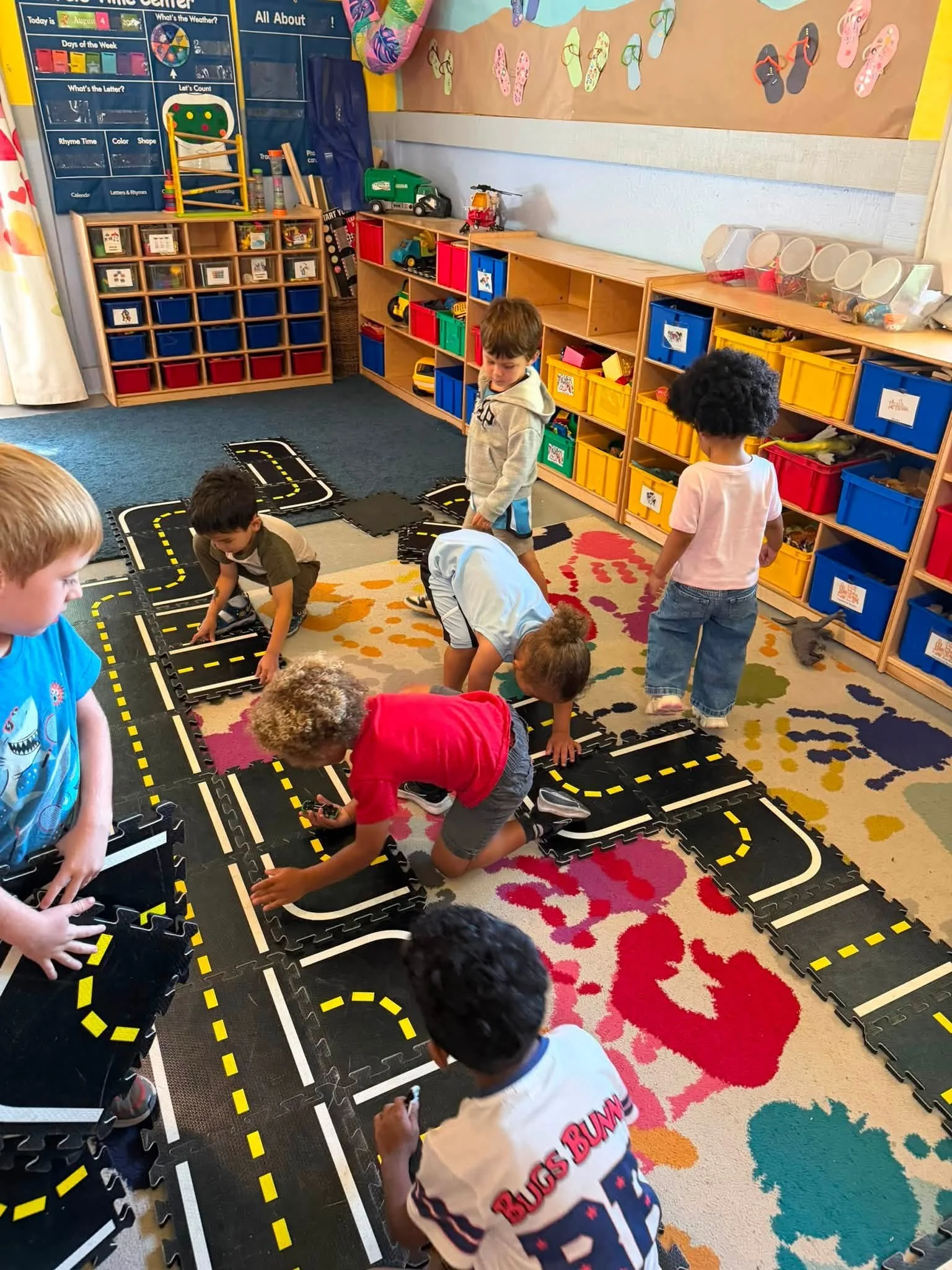 Children playing on a road-themed mat in a classroom, with toy vehicles and colorful storage bins on shelves, and decorations on the wall.
