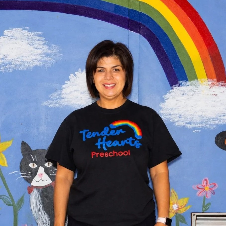 A teacher smiling in front of a colorful mural with a rainbow, clouds, a butterfly, a cat, and a ladybug. She is wearing a white T-shirt that says "Tender Hearts Preschool."