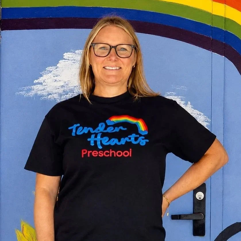 A teacher with long blonde hair smiling, standing in front of a colorful rainbow and clouds painted on a door.