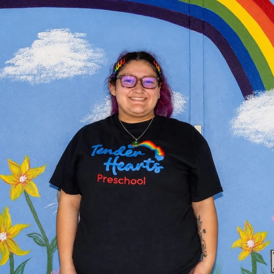 A smiling teacher with long curly hair standing in front of a blue wall painted with a colorful rainbow, flowers, clouds, a black cat, and an orange butterfly.